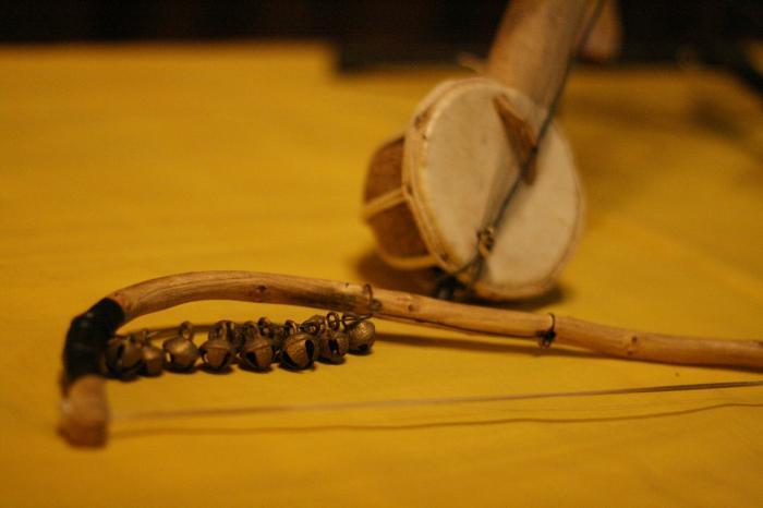 Un instrumento musical tradicional con un arco de madera y pequeñas campanas descansa sobre un paño amarillo. Detrás, un tambor con un cuerpo de cuerda añade un toque cultural.
