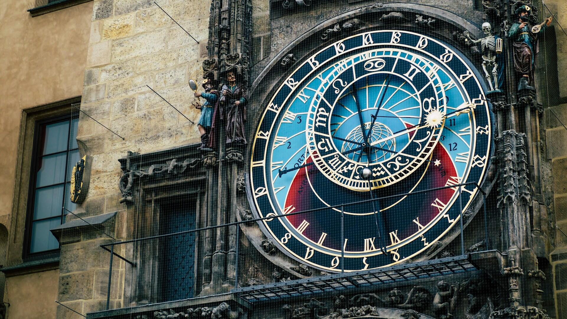 Reloj astronómico con diseños intrincados y símbolos del zodíaco, con estatuas junto a la esfera del reloj en un edificio antiguo.