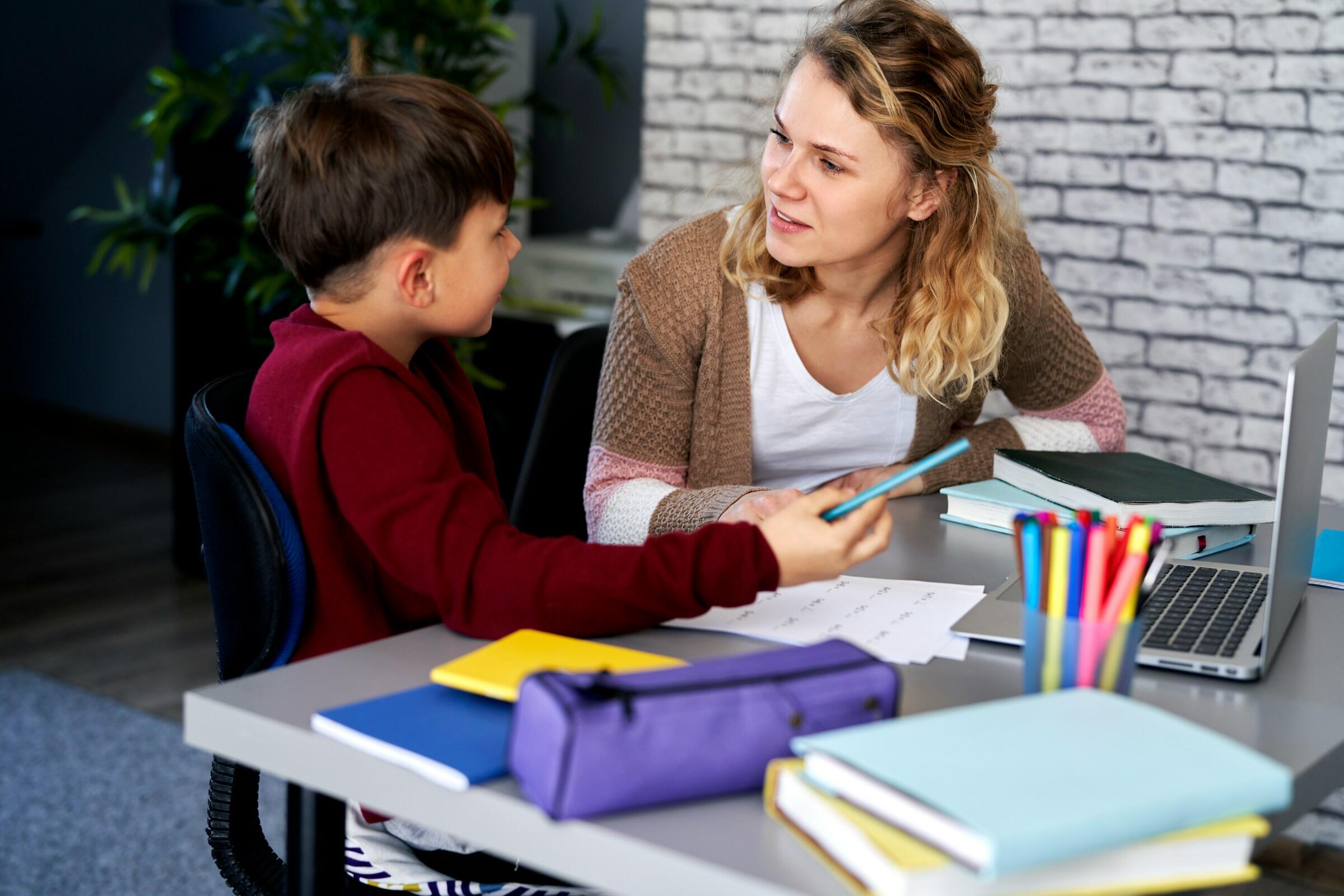 Un niño y un adulto están sentados a una mesa cubierta de libros, material de papelería y un ordenador portátil, conversando.
