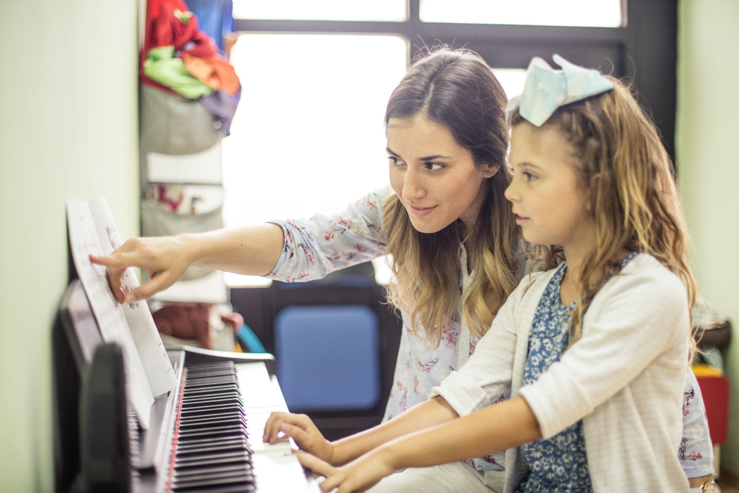 Una mujer señala una partitura mientras enseña a un niño a tocar el piano en un aula colorida y organizada.