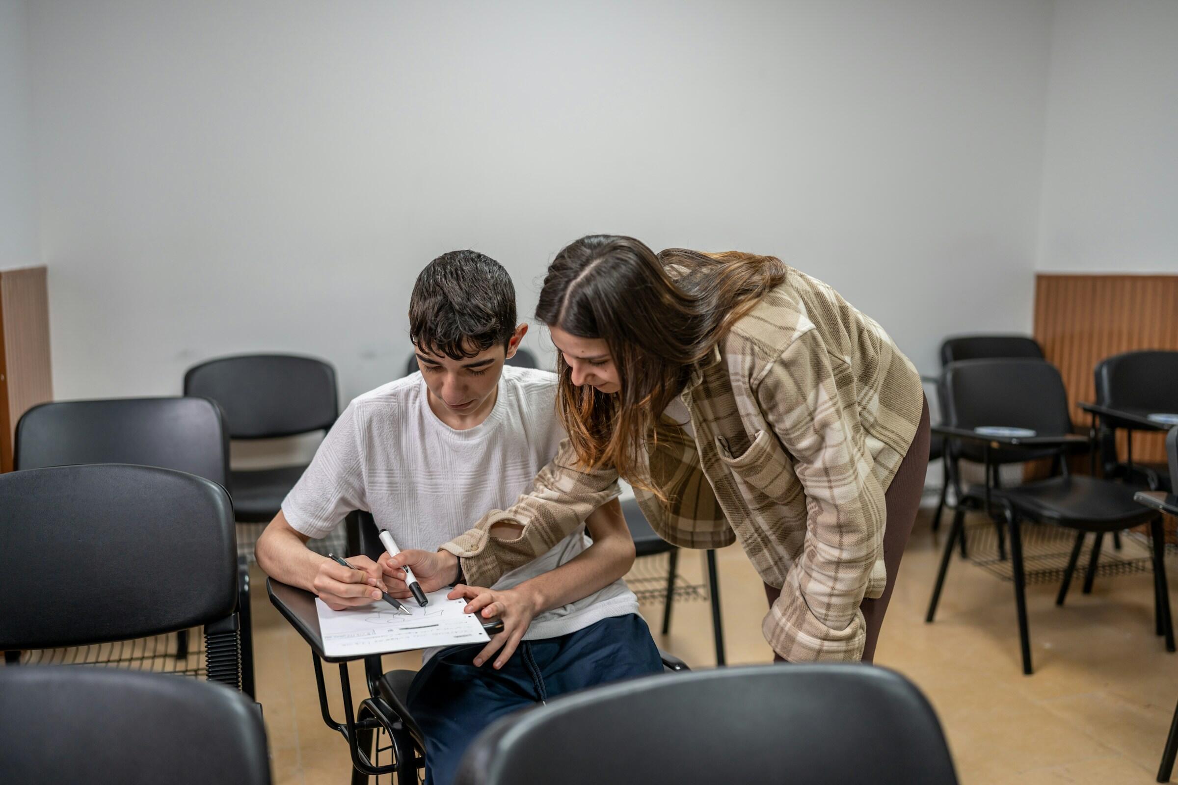 Una persona ayuda a otra en un aula, concentrándose en sus apuntes en un pupitre rodeado de sillas vacías.