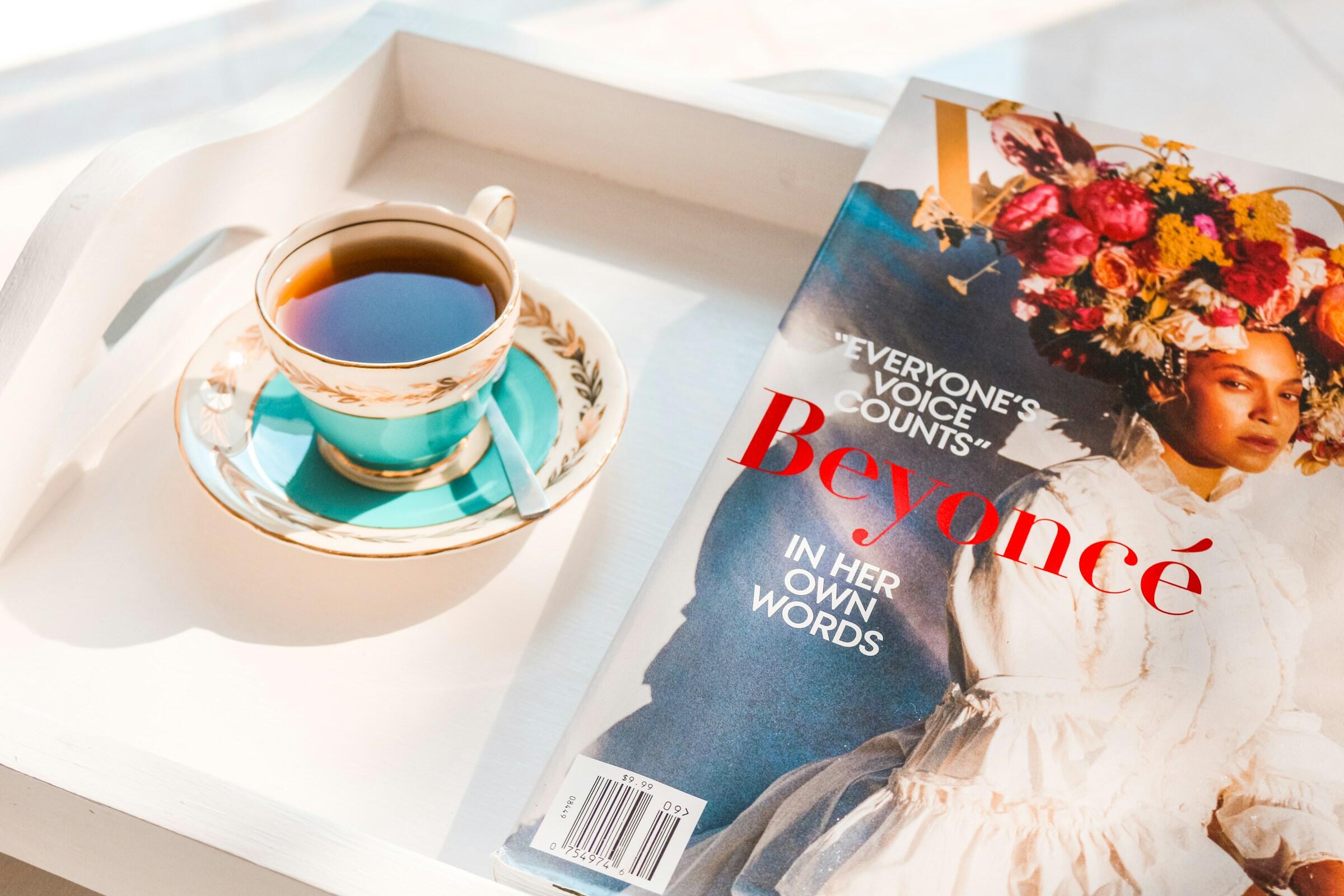 Una delicada taza de té sobre un platillo turquesa se encuentra junto a una revista en la que aparece un arreglo floral y el nombre «Beyoncé» destacado en un lugar prominente.