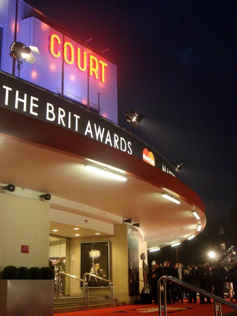 La entrada del recinto de los premios BRIT, con la palabra «COURT» en luces de neón, una alfombra roja y los asistentes bajo las luces nocturnas.