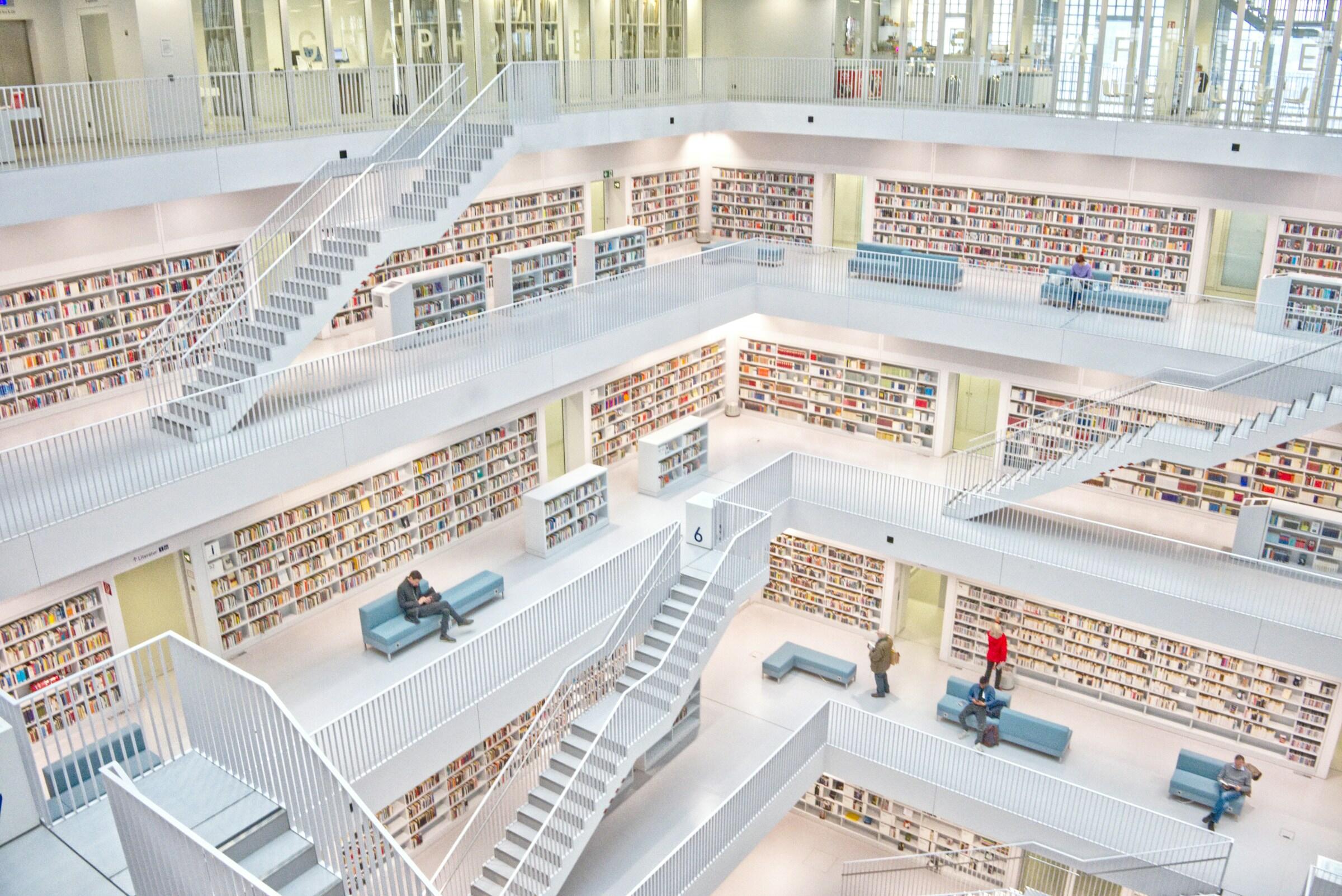El interior de una biblioteca moderna con varios niveles, paredes blancas y estanterías repletas de libros. La gente lee y camina por las escaleras abiertas.