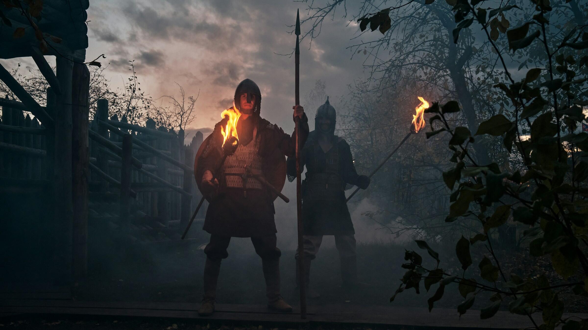 Dos figuras con armadura se encuentran en un entorno brumoso, sosteniendo lanzas y antorchas, con un bosque como telón de fondo bajo un cielo espectacular.