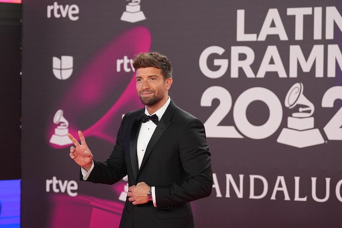 pablo alboran vestido con esmoquin posa en la alfombra roja de los Premios Grammy Latinos 2022, haciendo el signo de la paz frente a un fondo vibrante.
