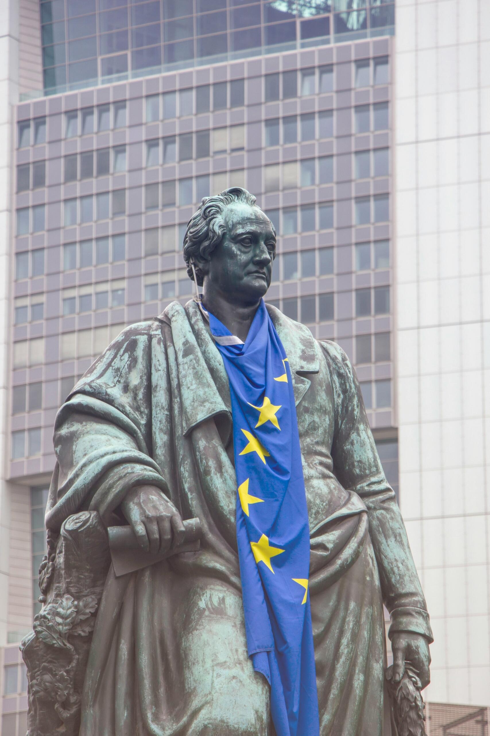 Una estatua de bronce de Goethe cubierta con una bandera azul de la Unión Europea, con un edificio moderno como telón de fondo.