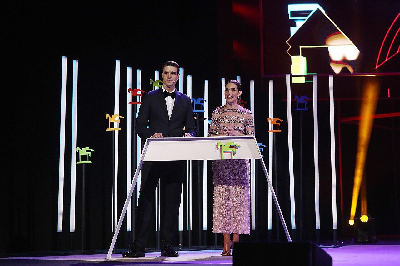 Un hombre con esmoquin y una mujer con un vestido brillante están de pie en el escenario de los premios ondas, rodeados de coloridas decoraciones iluminadas en el escenario.