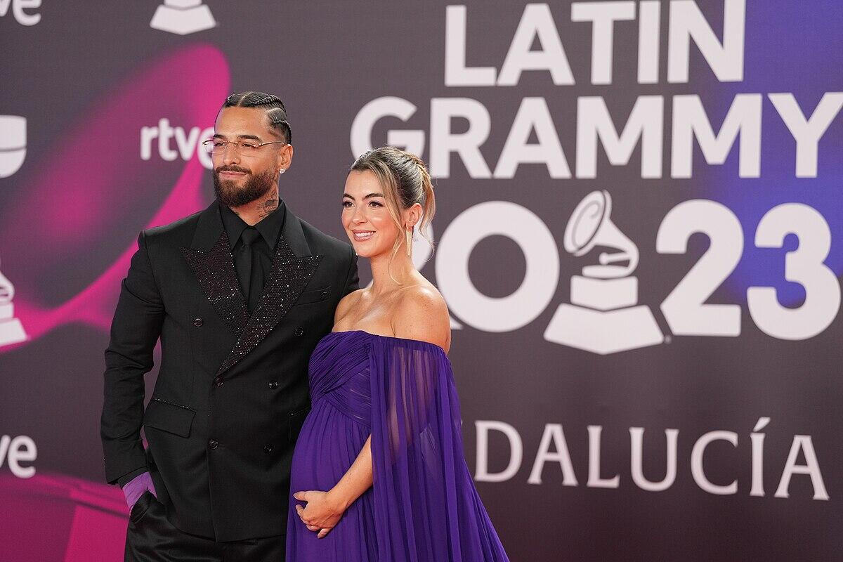 maluma y su pareja posan juntos en la alfombra roja de los Premios Grammy Latinos, luciendo elegantes trajes y con un fondo con la marca del evento.