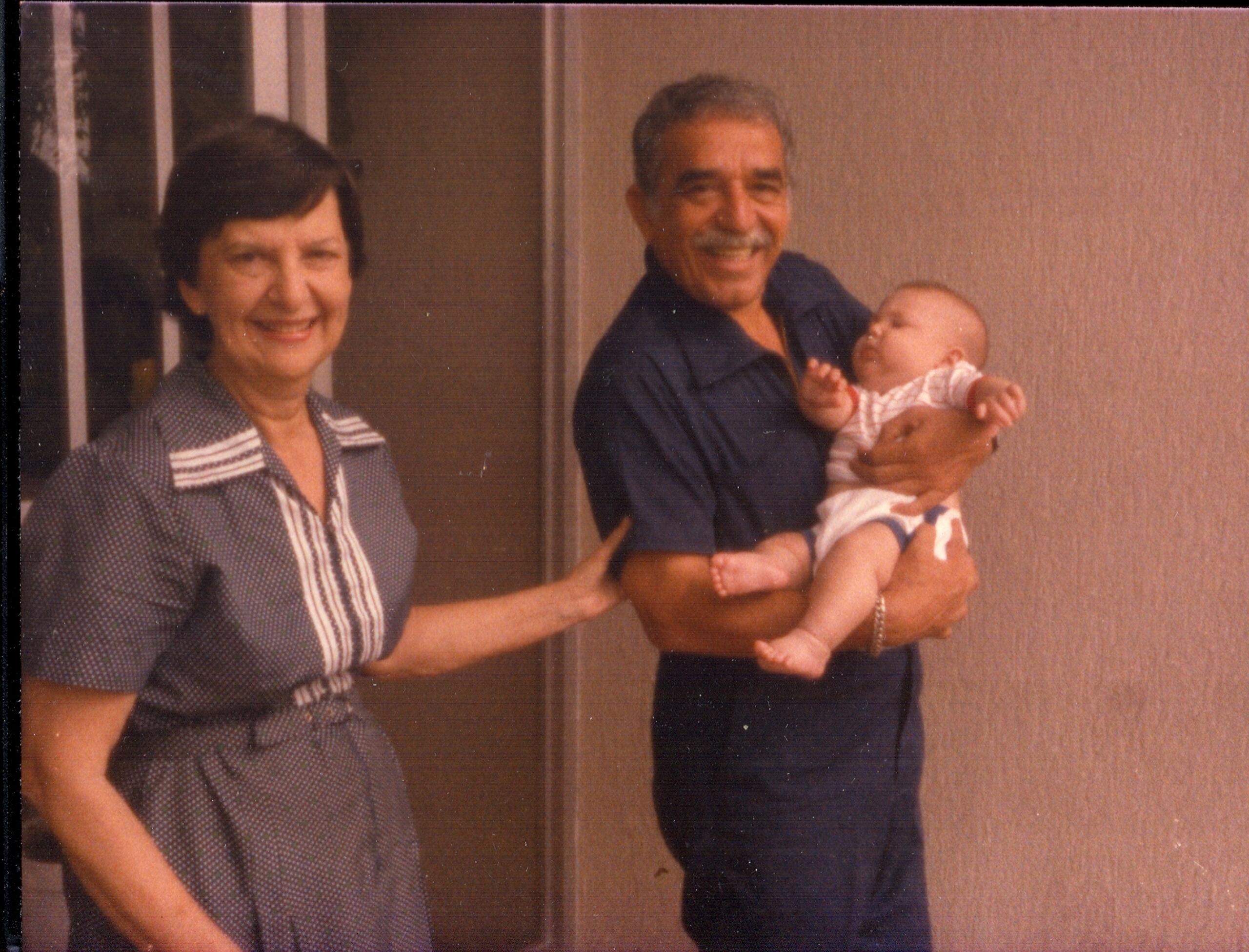 Un mujer y gabriel garcia marquez esta de pie, con el hombre sosteniendo a un bebé vestido con ropa blanca. Se encuentran en un ambiente informal en interiores.