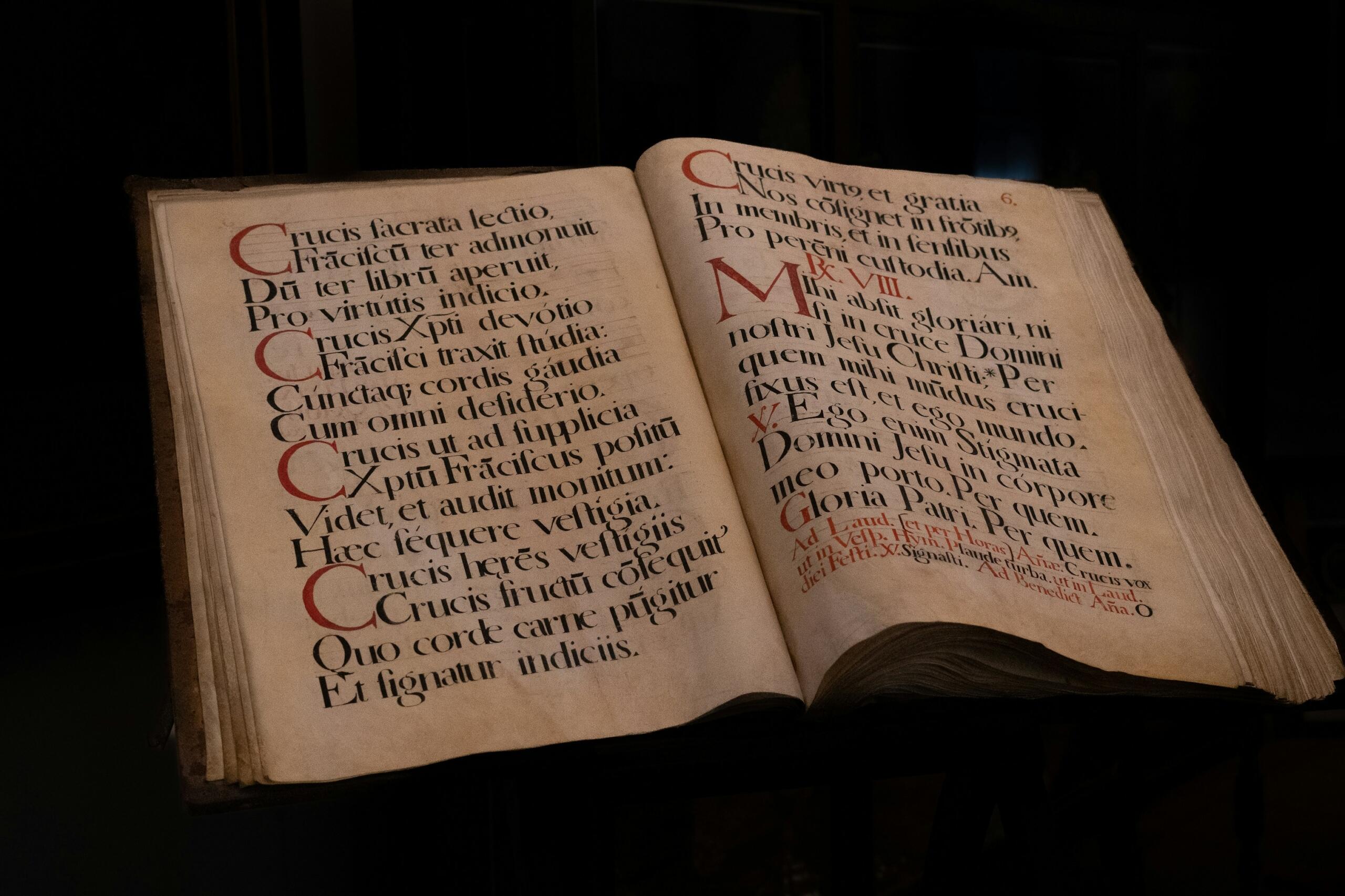 Un antiguo libro abierto muestra un texto iluminado en rojo y negro, con pasajes religiosos en latín y letras iniciales decorativas.
