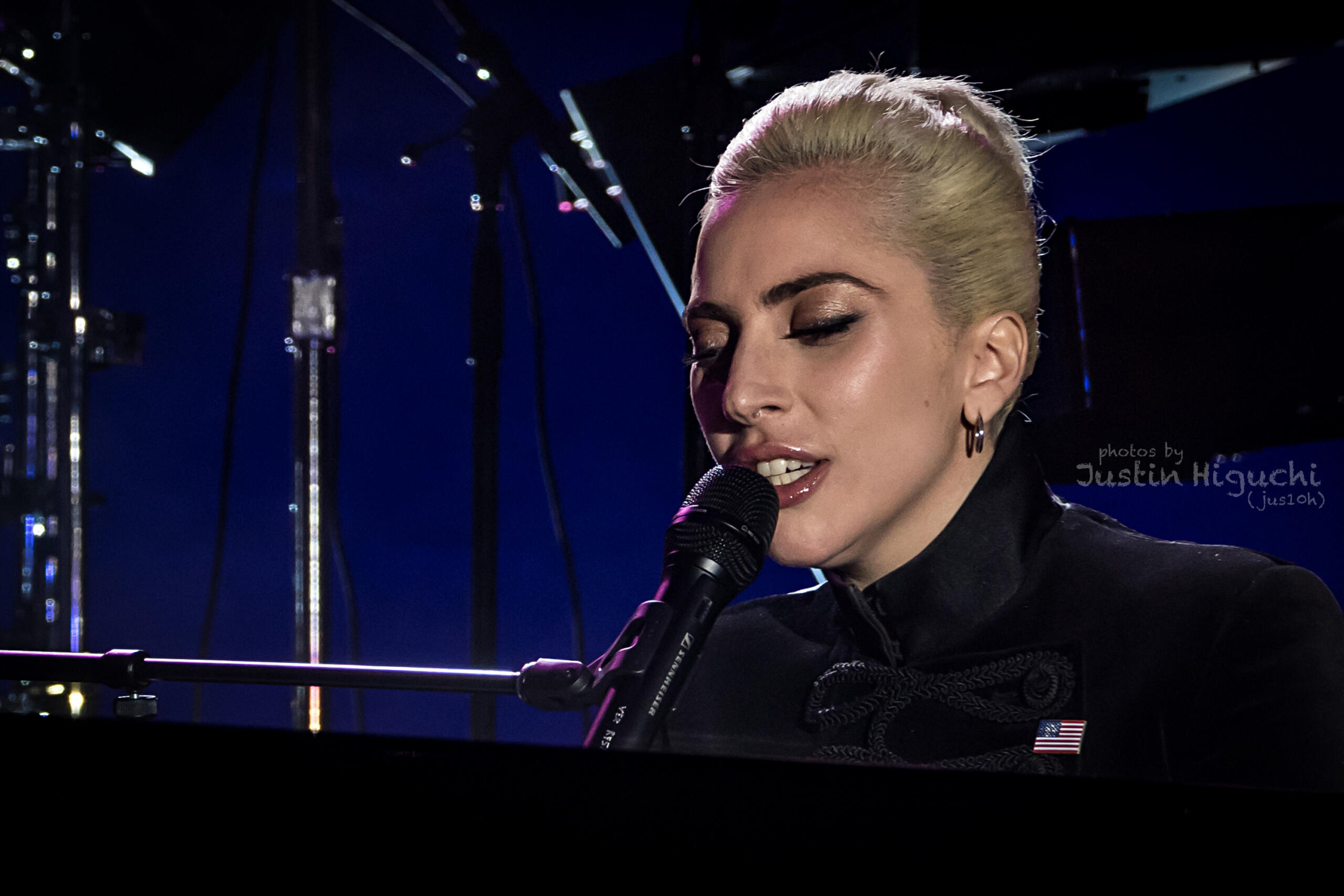 la artista lay gaga vestida con traje formal canta al micrófono mientras está sentado al piano, con un fondo azul oscuro y luces de escenario iluminando la escena.