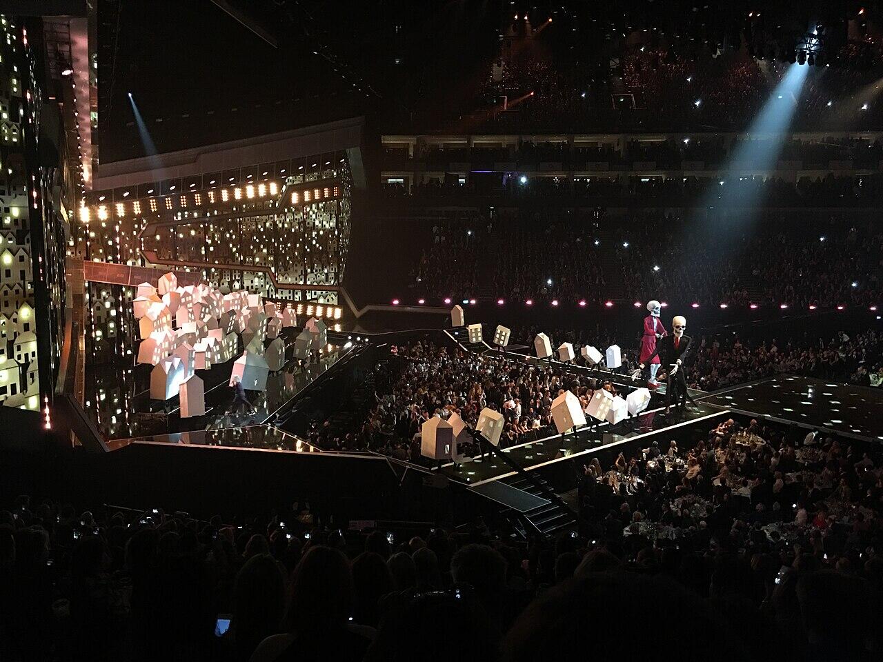 el escenario de los brit awards iluminado con luces, con artistas y público, decorado con elementos escenográficos y una gran multitud.