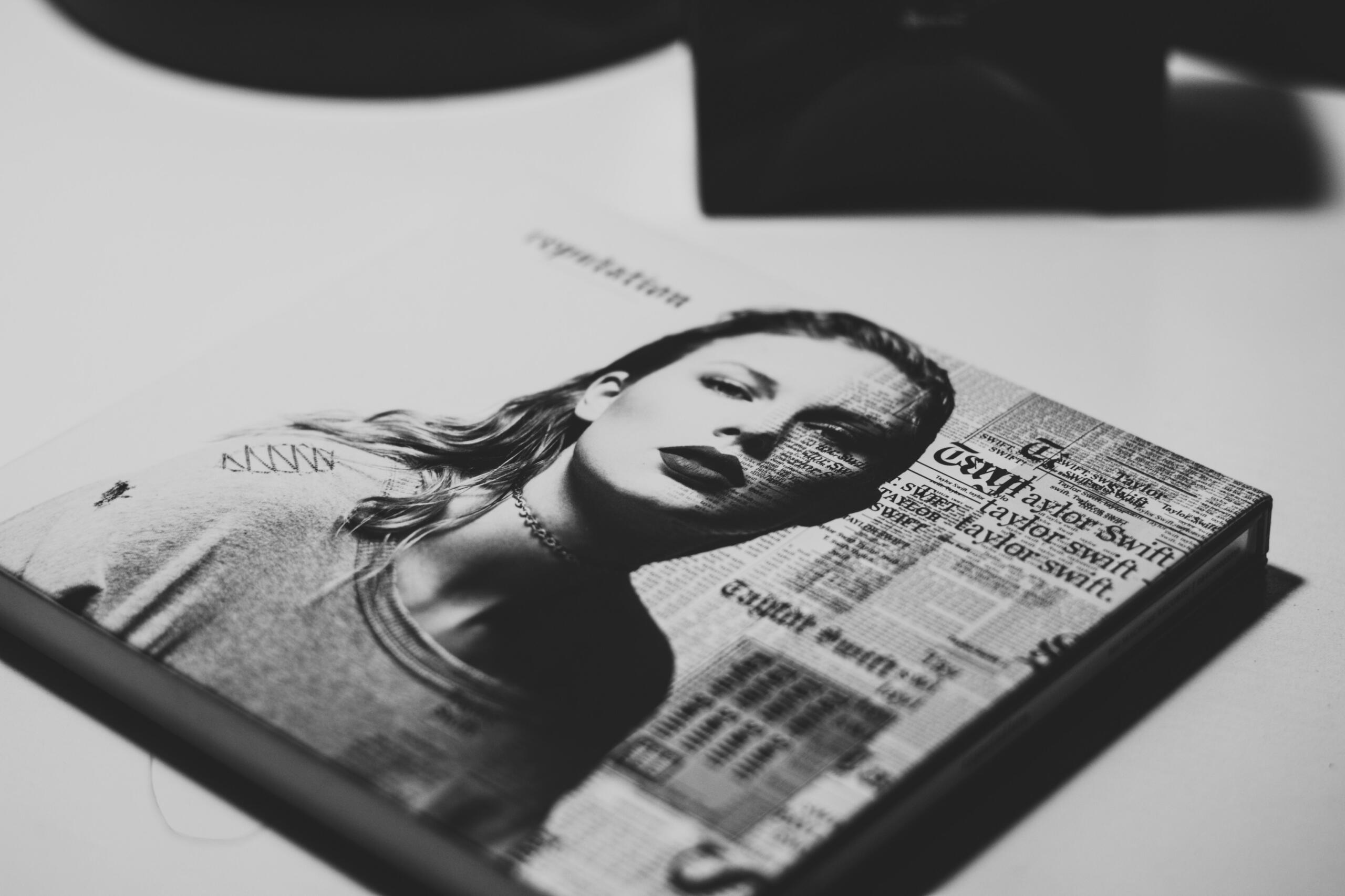 Primer plano de la portada de un álbum de taylor swift en la que aparece el retrato en blanco y negro de una mujer con texto y recortes de periódico superpuestos sobre su imagen.