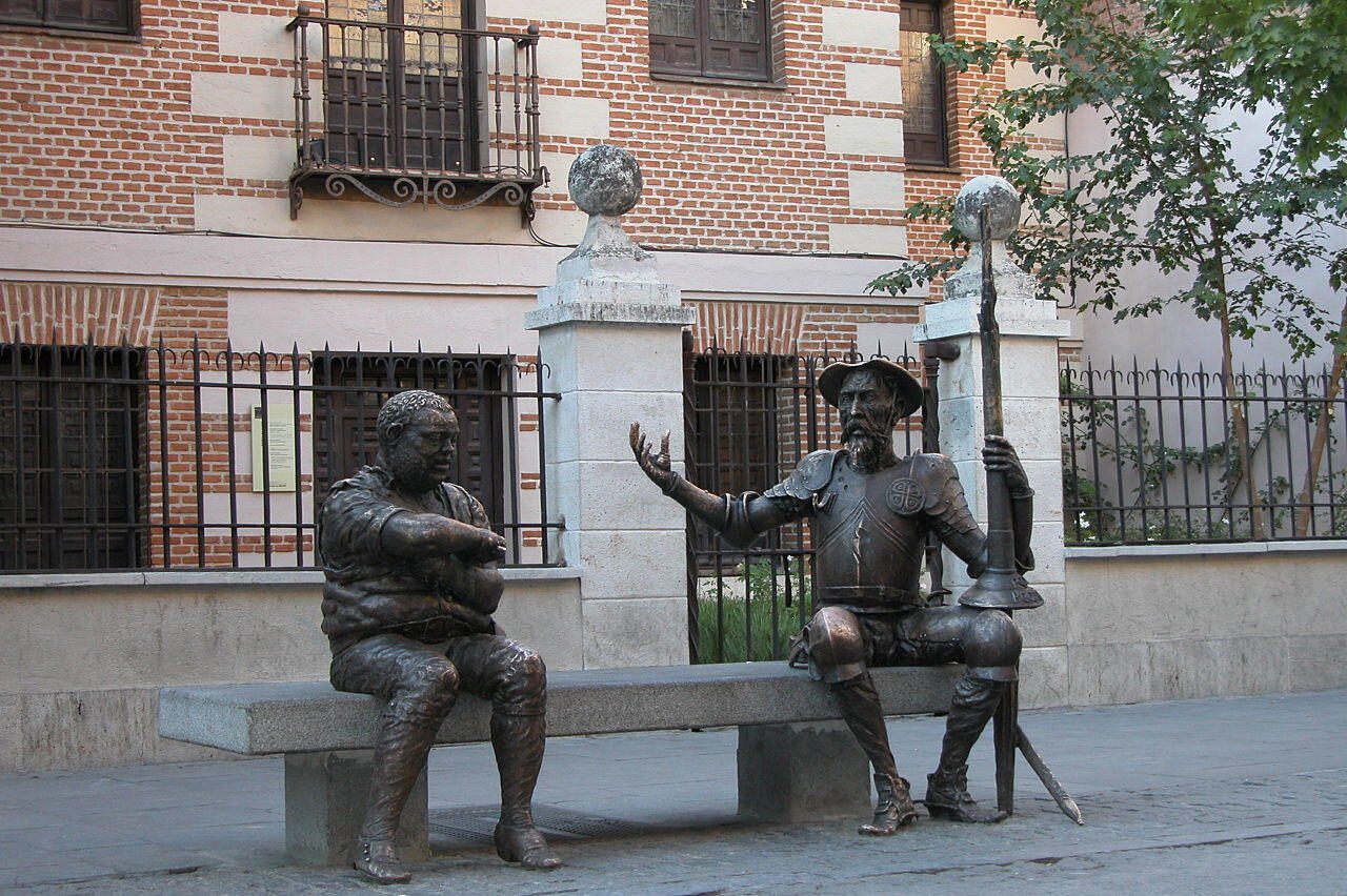 Dos estatuas de quijote y sancho panza en bronce se sientan en un banco, una con armadura haciendo un gesto y la otra con aspecto relajado, con un edificio de ladrillo como telón de fondo.