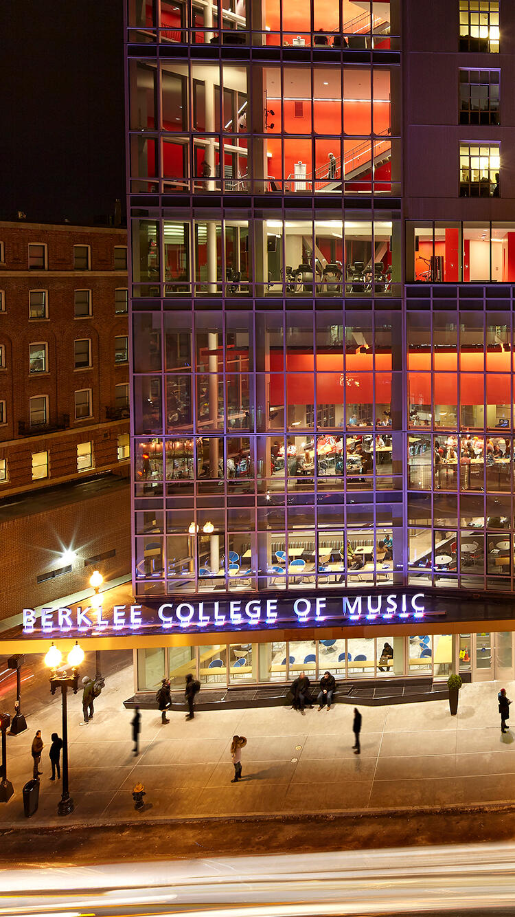 Vista nocturna del edificio del Berklee College of Music. La fachada de cristal brilla con la cálida luz roja del interior. La calle abajo está llena de estudiantes y luces.
