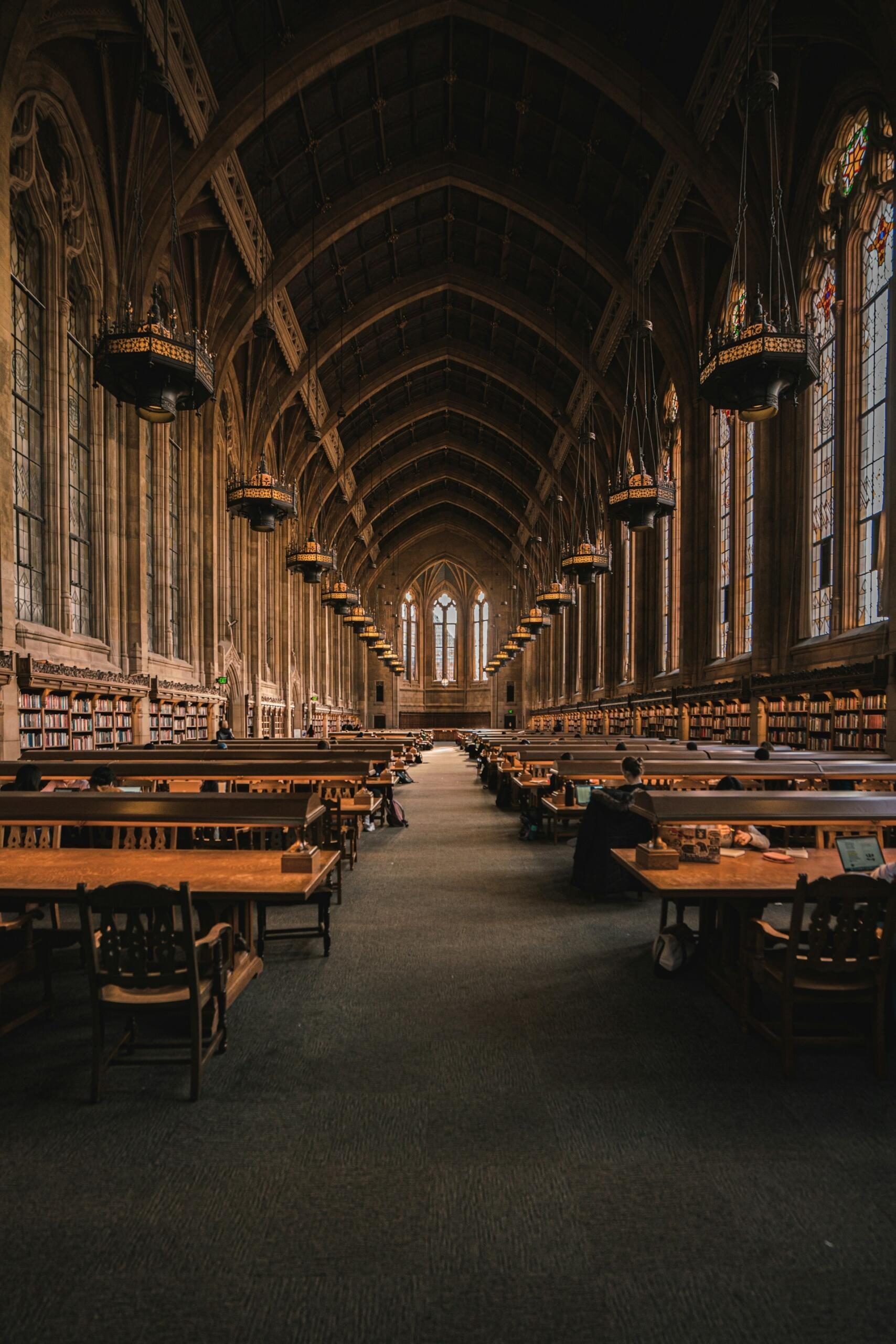 Interior de biblioteca de estilo gótico con altos techos abovedados y vidrieras. Mesas y estanterías de madera se alinean en la sala, creando un ambiente sereno y propicio para el estudio.A