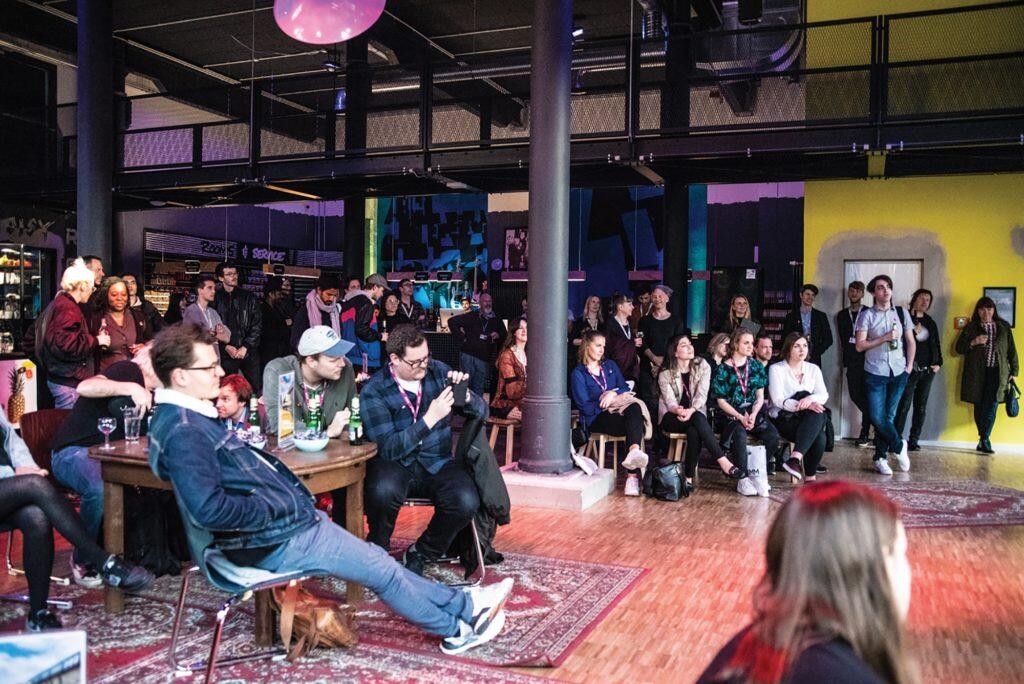 Un grupo diverso de personas está sentado y de pie en un animado local cubierto, escuchando con atención. La sala es vibrante, con paredes coloridas y una iluminación cálida.