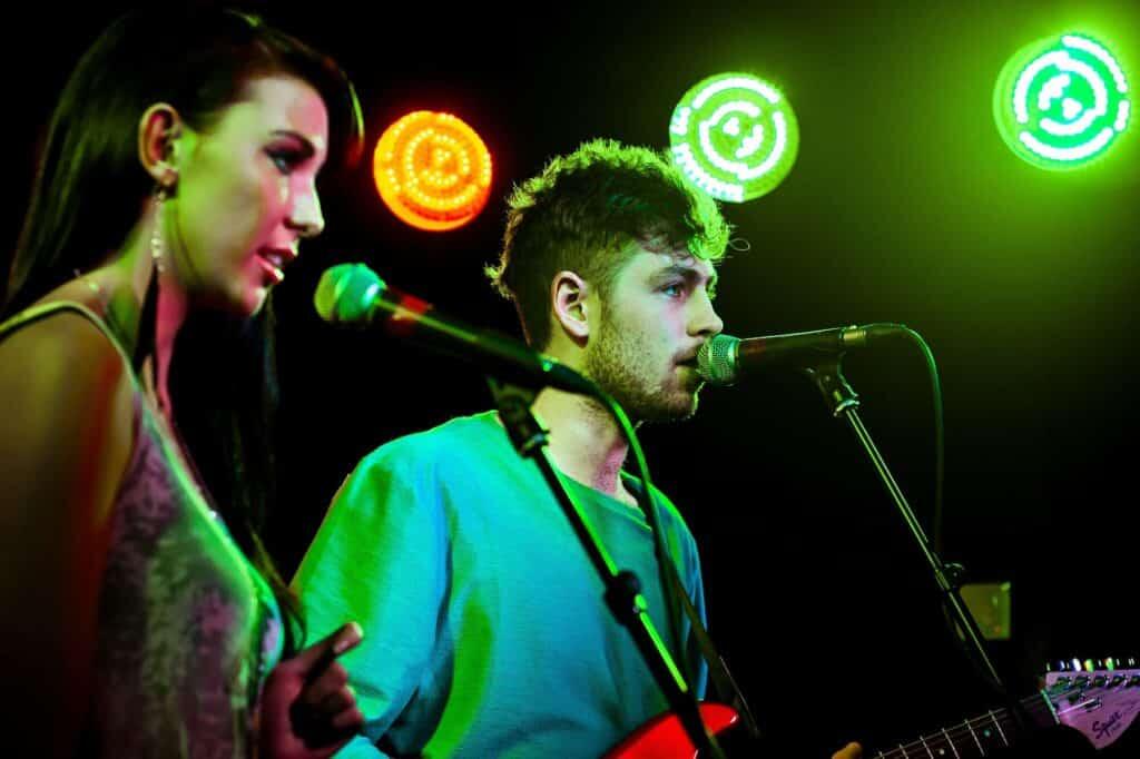 Un hombre y una mujer cantan ante los micrófonos en el escenario, iluminados por focos de colores verde y naranja. El ambiente es enérgico y concentrado.