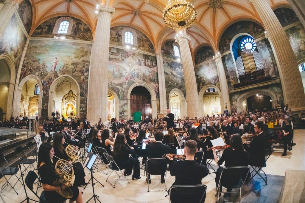 Una gran orquesta toca en una catedral ornamentada, con grandes pilares, murales y una lámpara de araña. El público escucha con atención, creando un ambiente elegante.