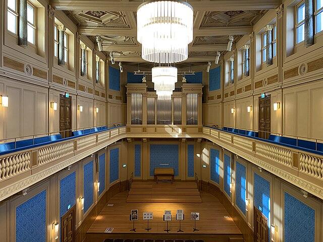 Lujoso interior de sala de conciertos con paredes azules, balcón ornamentado y grandes candelabros. El escenario de madera y el órgano crean un ambiente elegante y refinado.