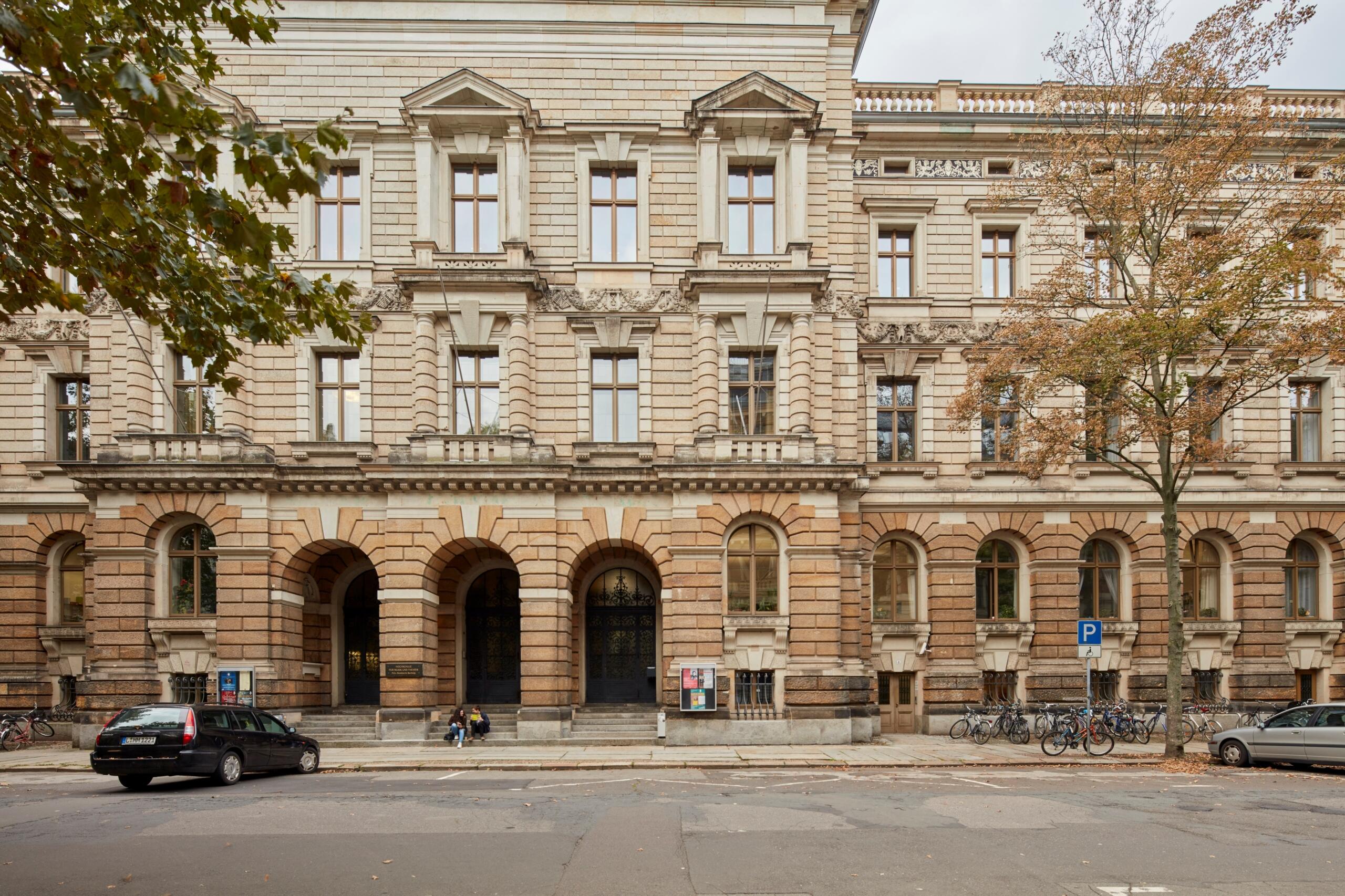Fachada de edificio histórico con entrada en arco y ventanas simétricas. Dos coches aparcados, bicicletas alineadas y árboles otoñales que añaden encanto al paisaje urbano.