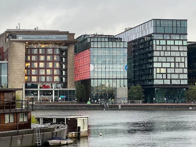 Los edificios modernos se alinean a lo largo de la ribera, con una prominente fachada acristalada en la que se lee «Conservatorium van Amsterdam». La calma del río y el cielo nublado crean un ambiente sereno.