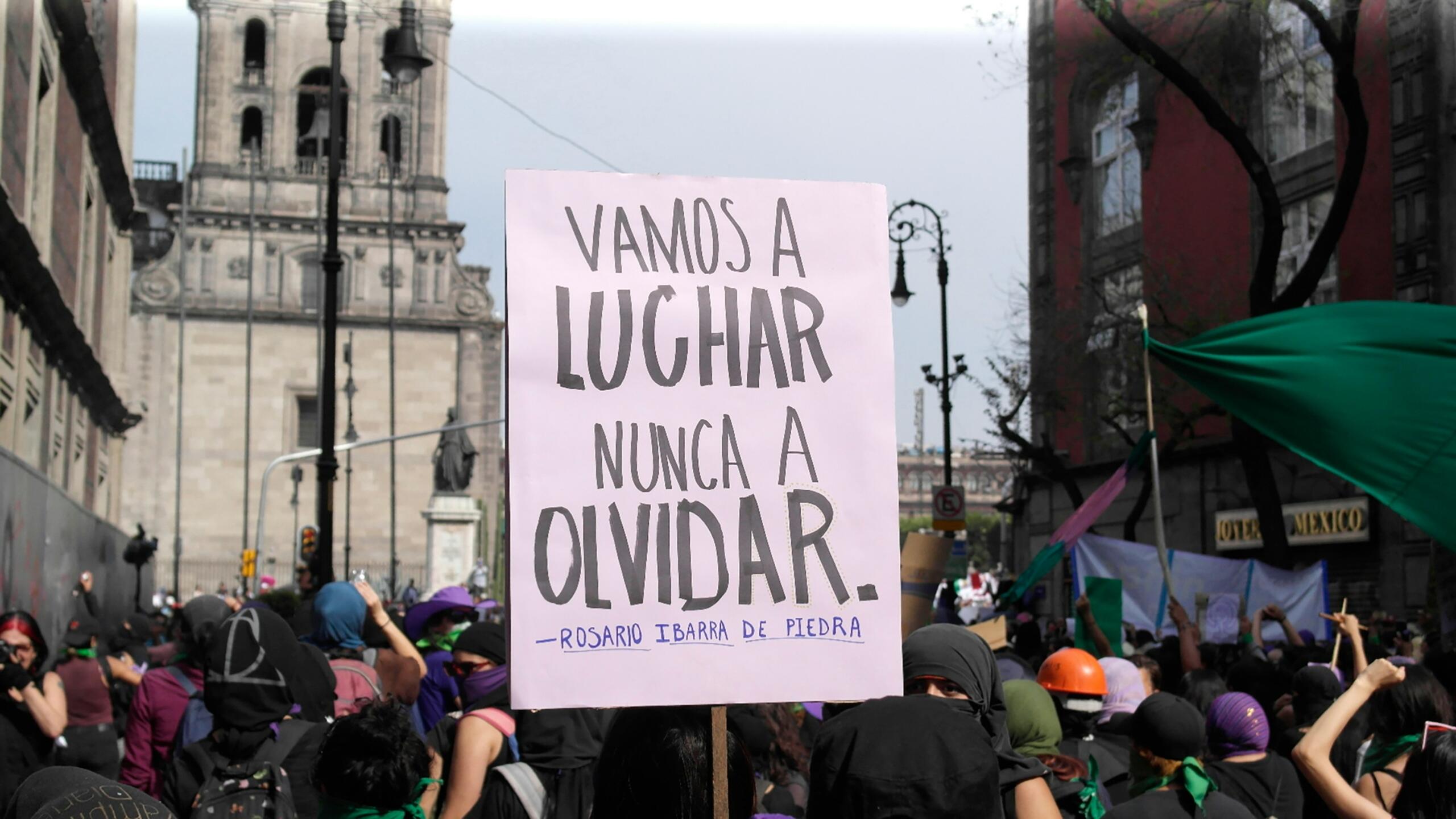 Una multitud marcha en protesta, sosteniendo un cartel que dice «Vamos a luchar, nunca a olvidar», cerca de un edificio histórico.
