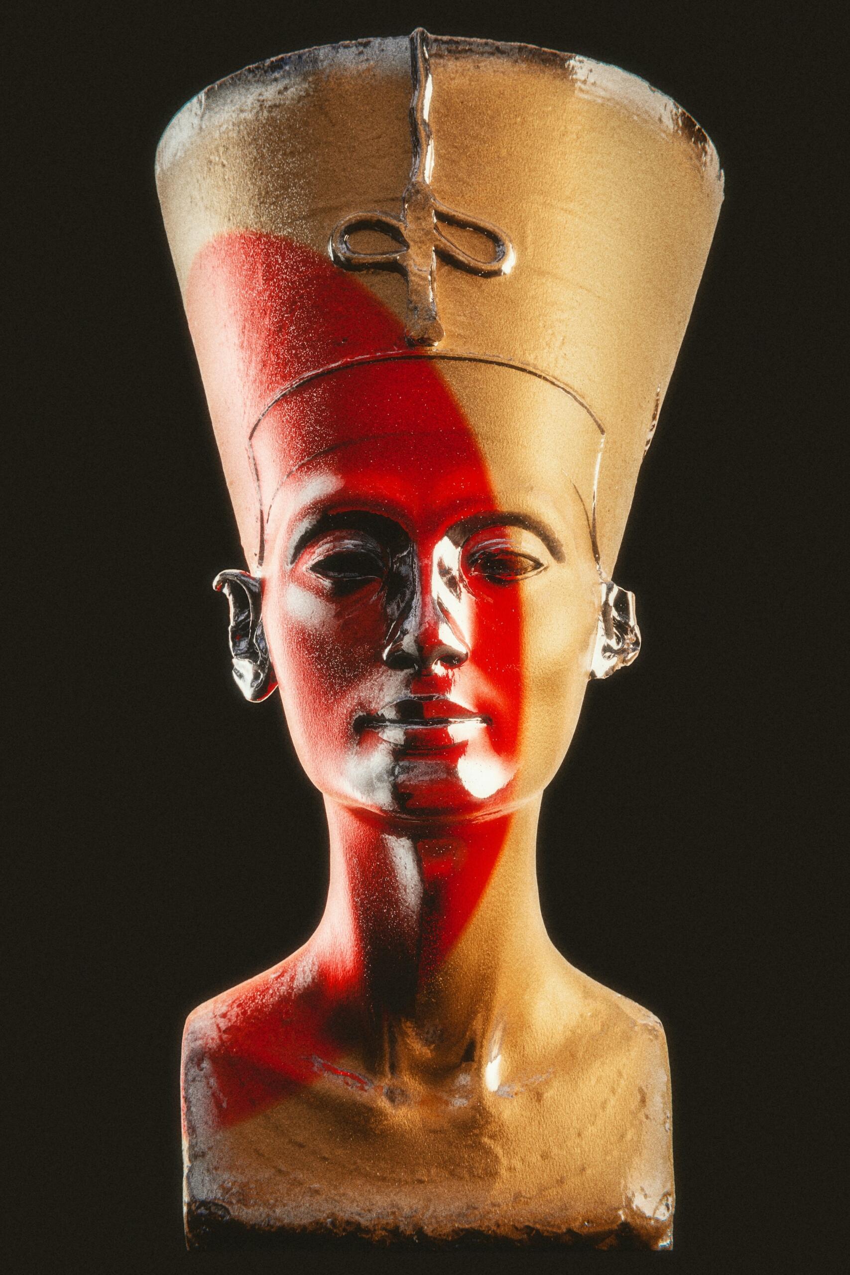 Una escultura de nefertiti detallada de un busto con una corona dorada, con detalles en rojo y negro en el cuello y los hombros, sobre un fondo oscuro.