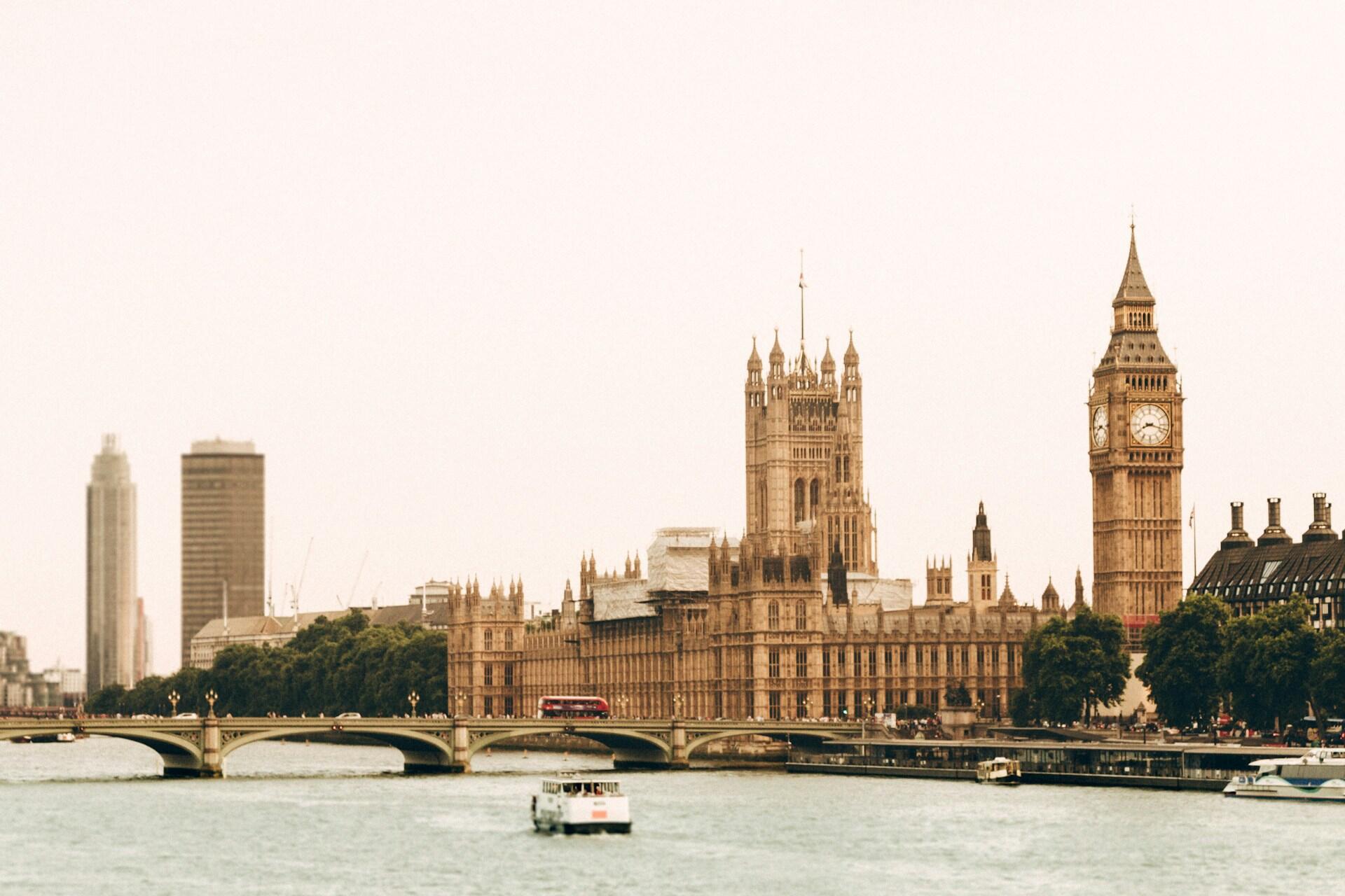 Vista emblemática del Parlamento y el Big Ben de Londres junto al río Támesis, bajo un cielo brumoso. Un autobús rojo de dos pisos cruza el puente de Westminster.