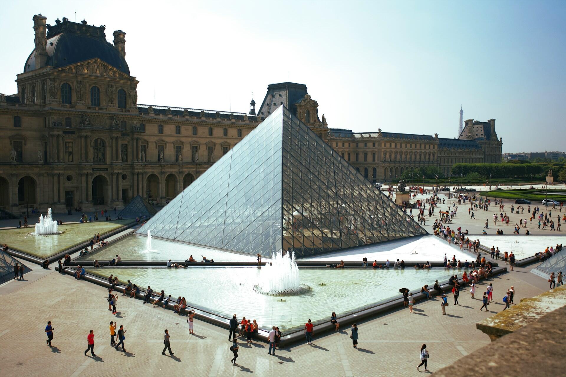 La gran pirámide de cristal del Louvre se alza en el patio del Louvre, rodeada de fuentes y gente. Los edificios históricos del palacio flanquean la escena bajo un cielo azul claro.