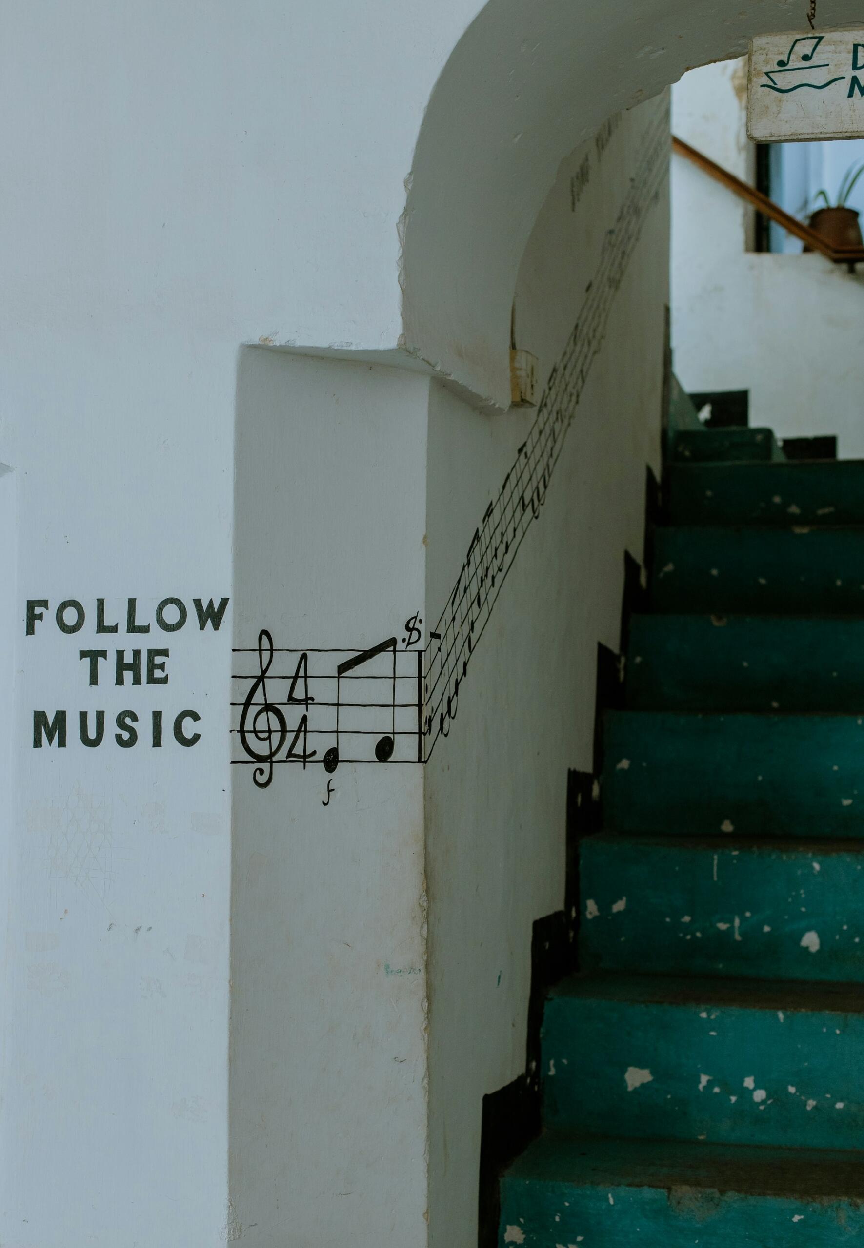 Una escalera verde azulada conduce hacia arriba con notas musicales pintadas en la pared junto a ella, acompañadas de la frase «Sigue la música». La escena resulta artística y acogedora.