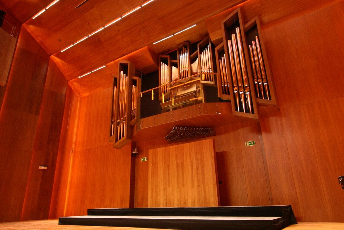 Un gran órgano de tubos está montado en una pared de madera en una sala de conciertos. Los altos tubos y los cálidos tonos de la madera transmiten una sensación de grandeza y elegancia.