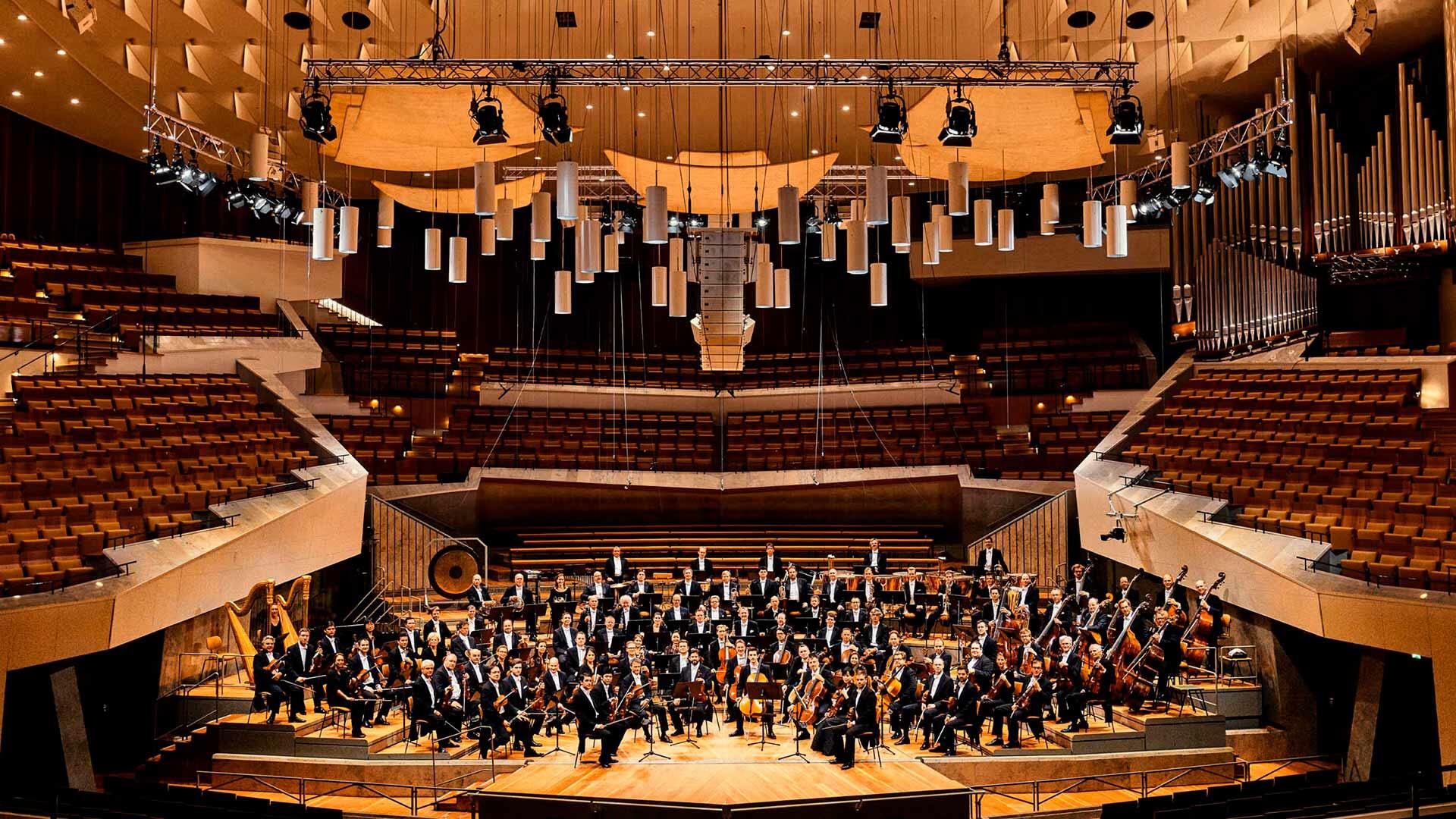 Una orquesta sinfónica actúa en un gran escenario en una amplia sala de conciertos, con un diseño moderno en gradas y una iluminación cálida, creando un ambiente elegante.