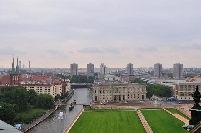 Vista aérea de un paisaje urbano con un río bordeado por edificios históricos y rascacielos modernos. En primer plano, bajo un cielo nublado, se extienden exuberantes prados verdes.