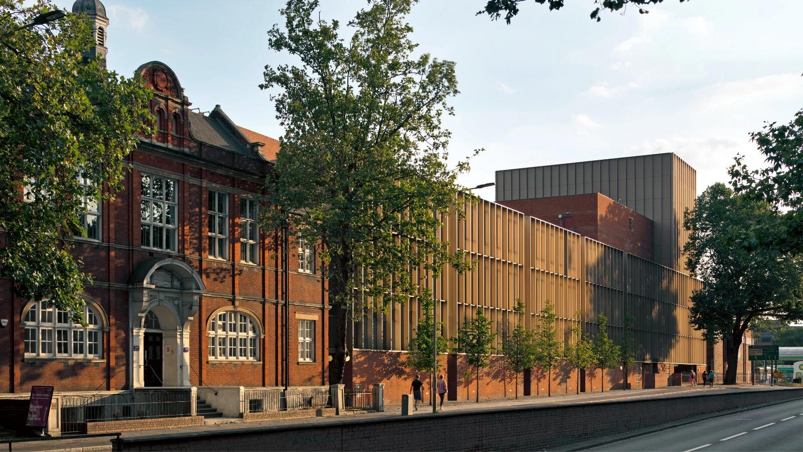 Vista de la calle de un moderno edificio de ladrillo con una fachada clásica a la izquierda, bordeada de árboles. La cálida luz del sol crea un ambiente tranquilo y acogedor.