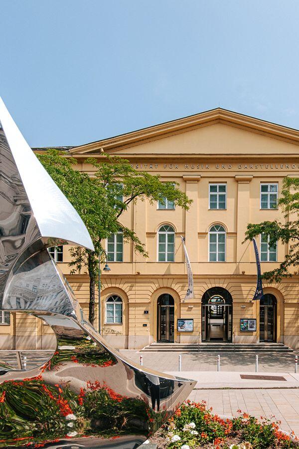 Edificio histórico de color beige con ventanas y entrada en arco, flanqueado por banderas. Una escultura metálica reflectante y flores vibrantes en primer plano. Cielo azul claro.