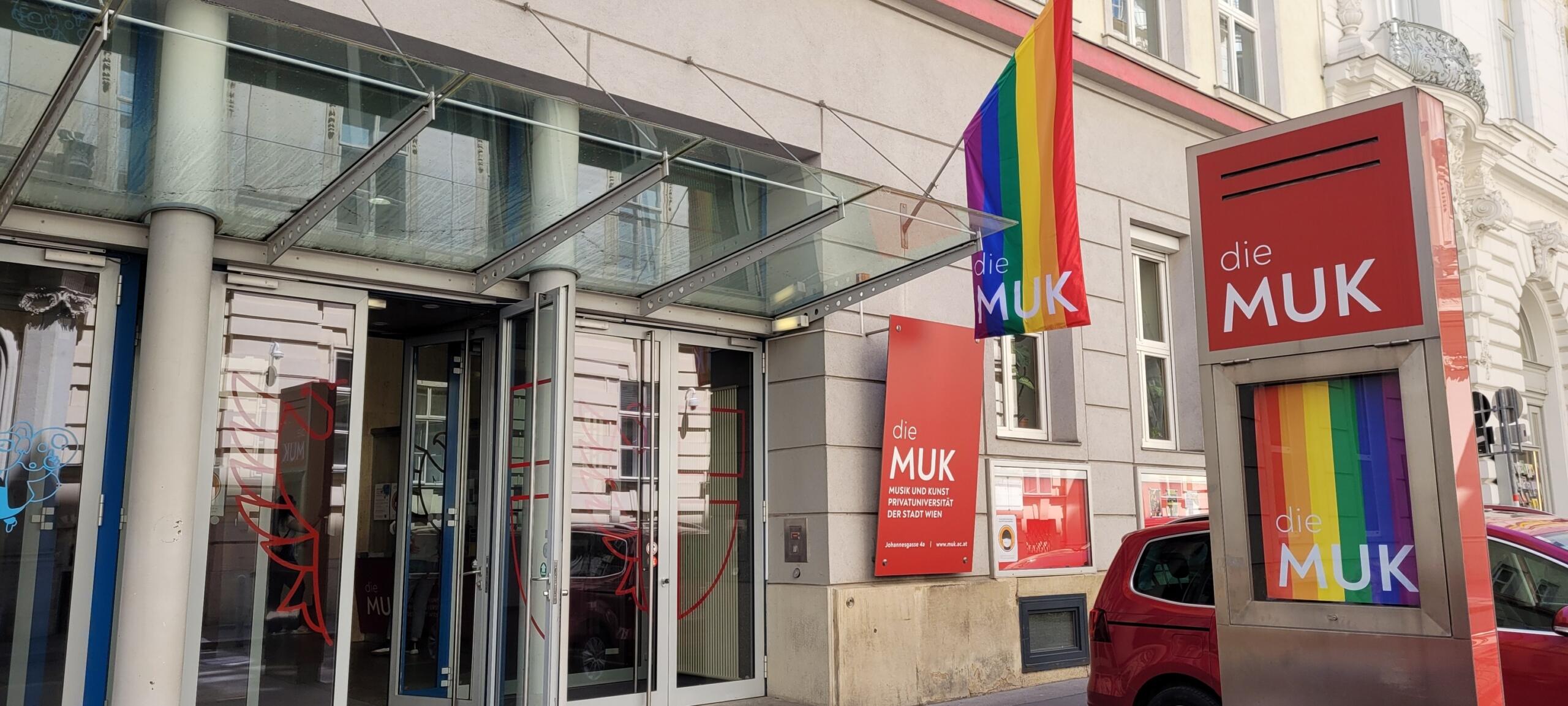 Entrada de «die MUK», con puertas de cristal, banderas arcoíris y letreros rojos con texto blanco bajo una marquesina transparente. Urbana y acogedora.