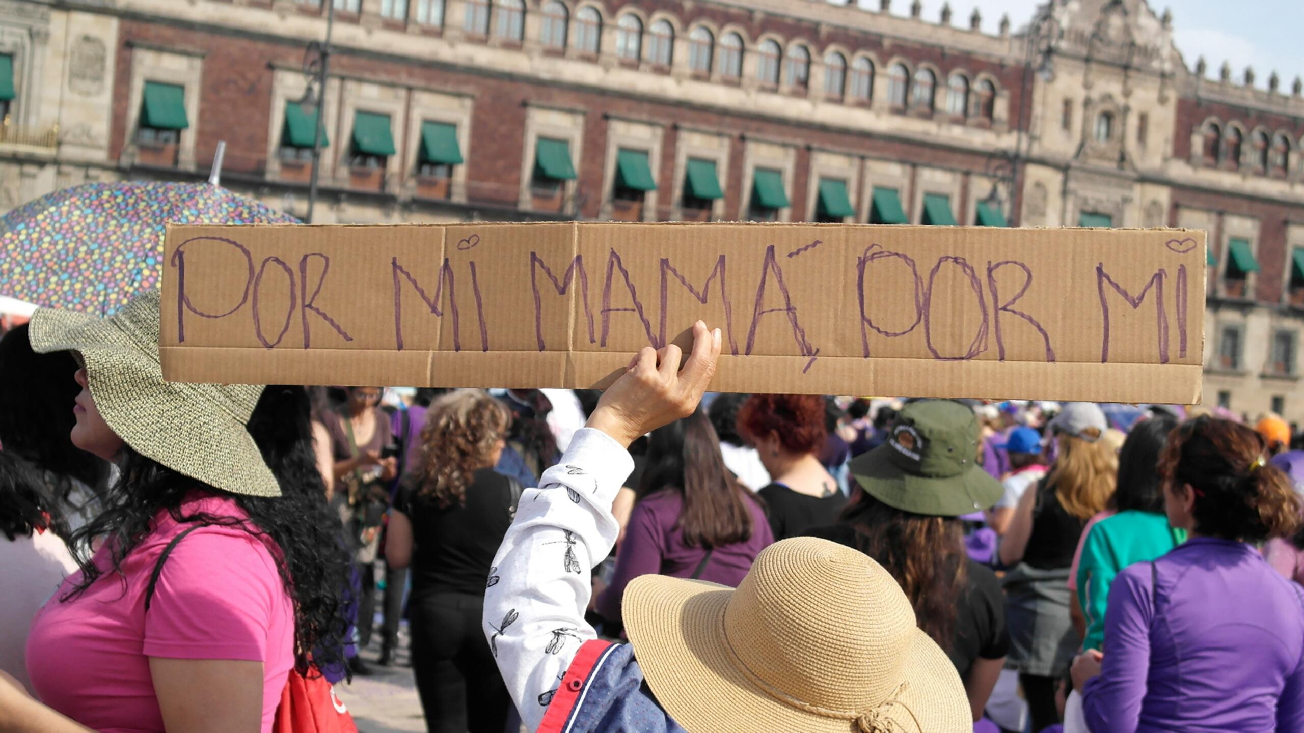 Una persona sostiene un cartel de cartón en el que se lee «Por mi mamá» en un concurrido evento al aire libre, con asistentes vestidos de morado.