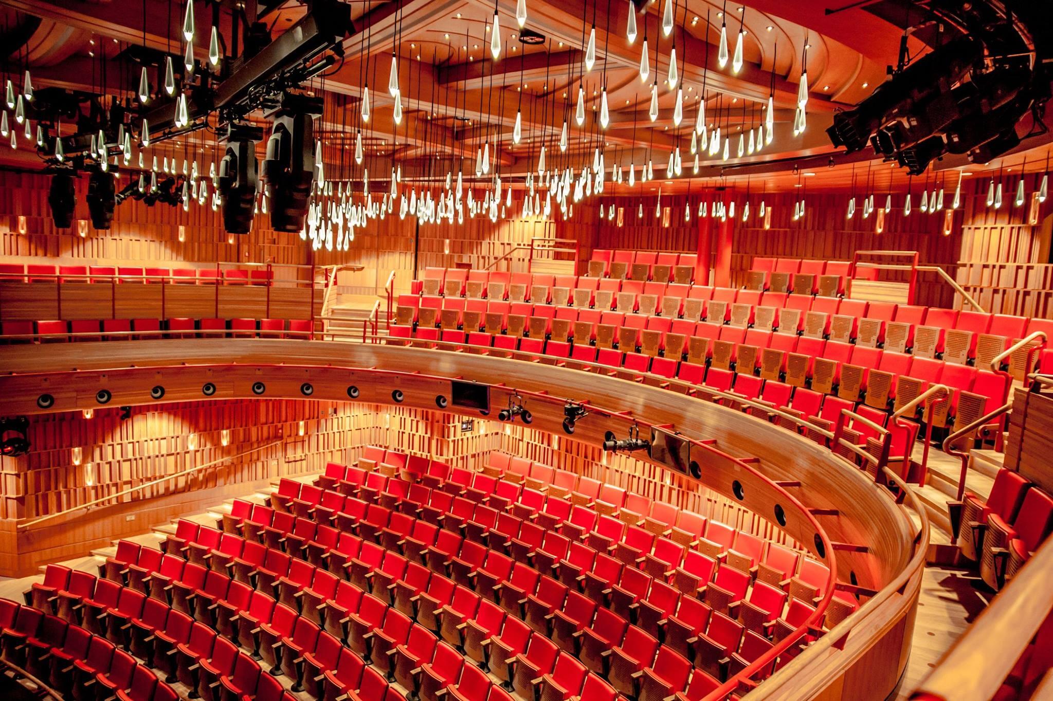 Elegante interior de teatro con butacas rojas y paredes curvas de madera. Modernas lámparas en forma de lágrima cuelgan del techo, creando un ambiente cálido.