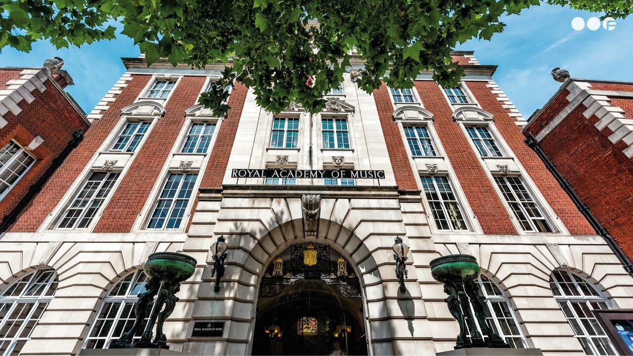 Edificio histórico con un gran arco con la inscripción «Real Academia de Música». Fachada de ladrillo rojo, ventanas altas y ramas frondosas bajo un cielo azul.