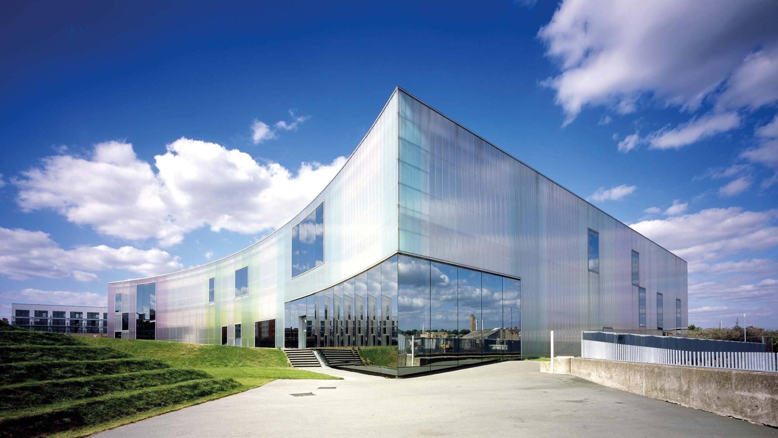 Moderno edificio acristalado con paneles reflectantes bajo un cielo azul brillante, rodeado de césped verde. La arquitectura es elegante y contemporánea, y transmite innovación.