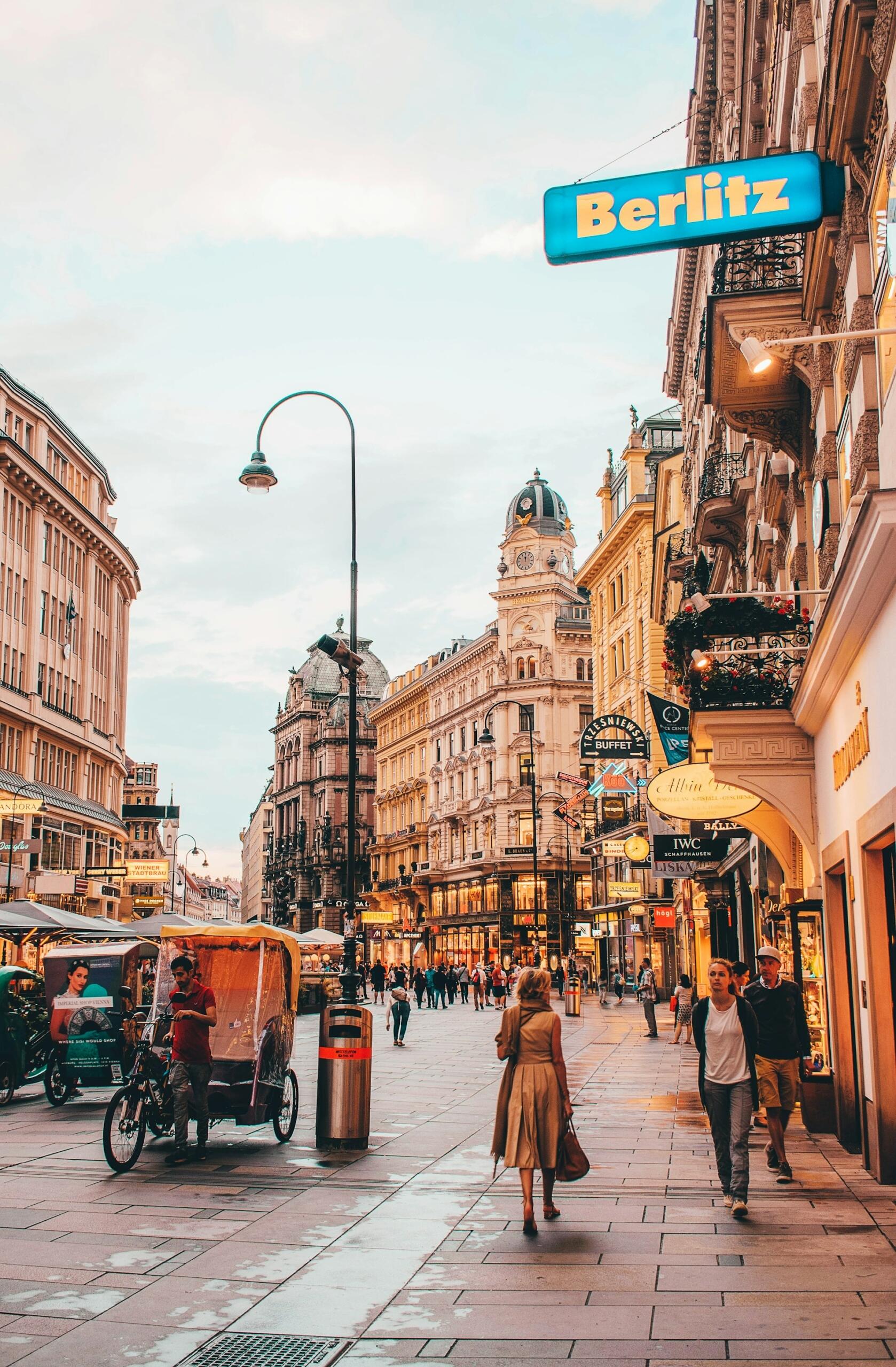 Calle bulliciosa de la ciudad al atardecer con arquitectura histórica. Gente caminando, un ciclista y un luminoso letrero de «Berlitz». Ambiente cálido y animado.