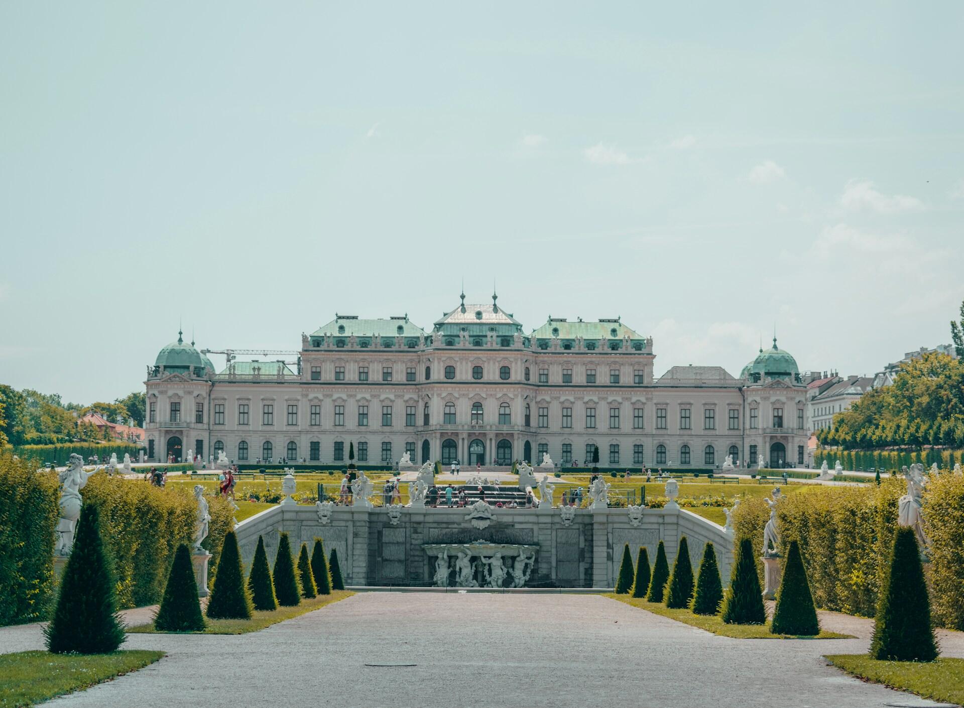 Elegante palacio barroco con un techo abovedado verde, rodeado de cuidados jardines y estatuas, bajo un cielo azul claro, que transmite grandeza y serenidad.