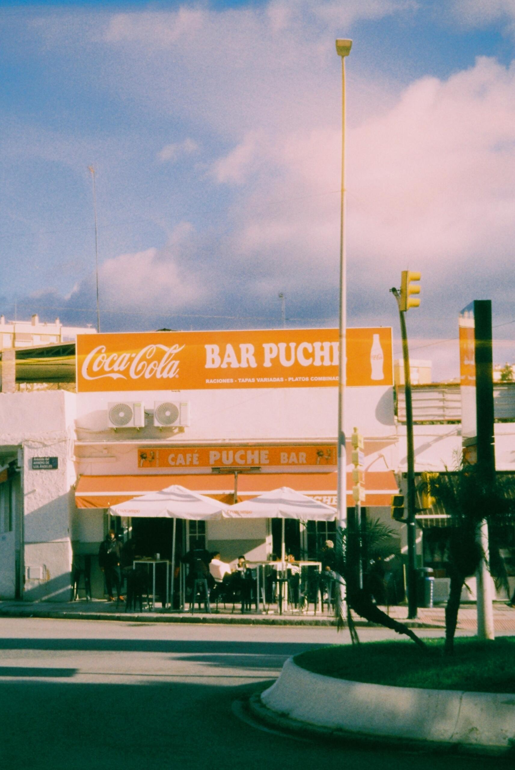 Vista de la calle del «Bar Puche», con un cartel de Coca-Cola. Se ve una terraza con sombrillas blancas bajo un cielo azul salpicado de nubes esponjosas.