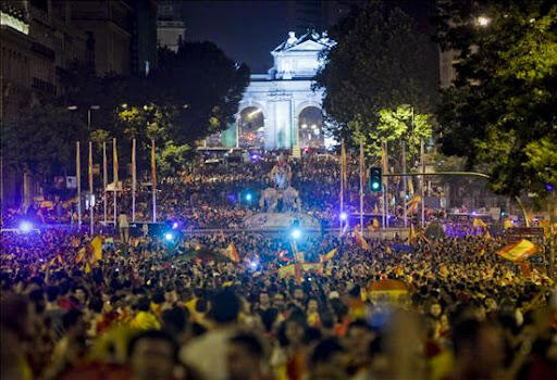 Una multitud celebrando por la noche cerca de un monumento brillantemente iluminado, ondeando banderas bajo los árboles y las farolas, en un ambiente festivo y animado.