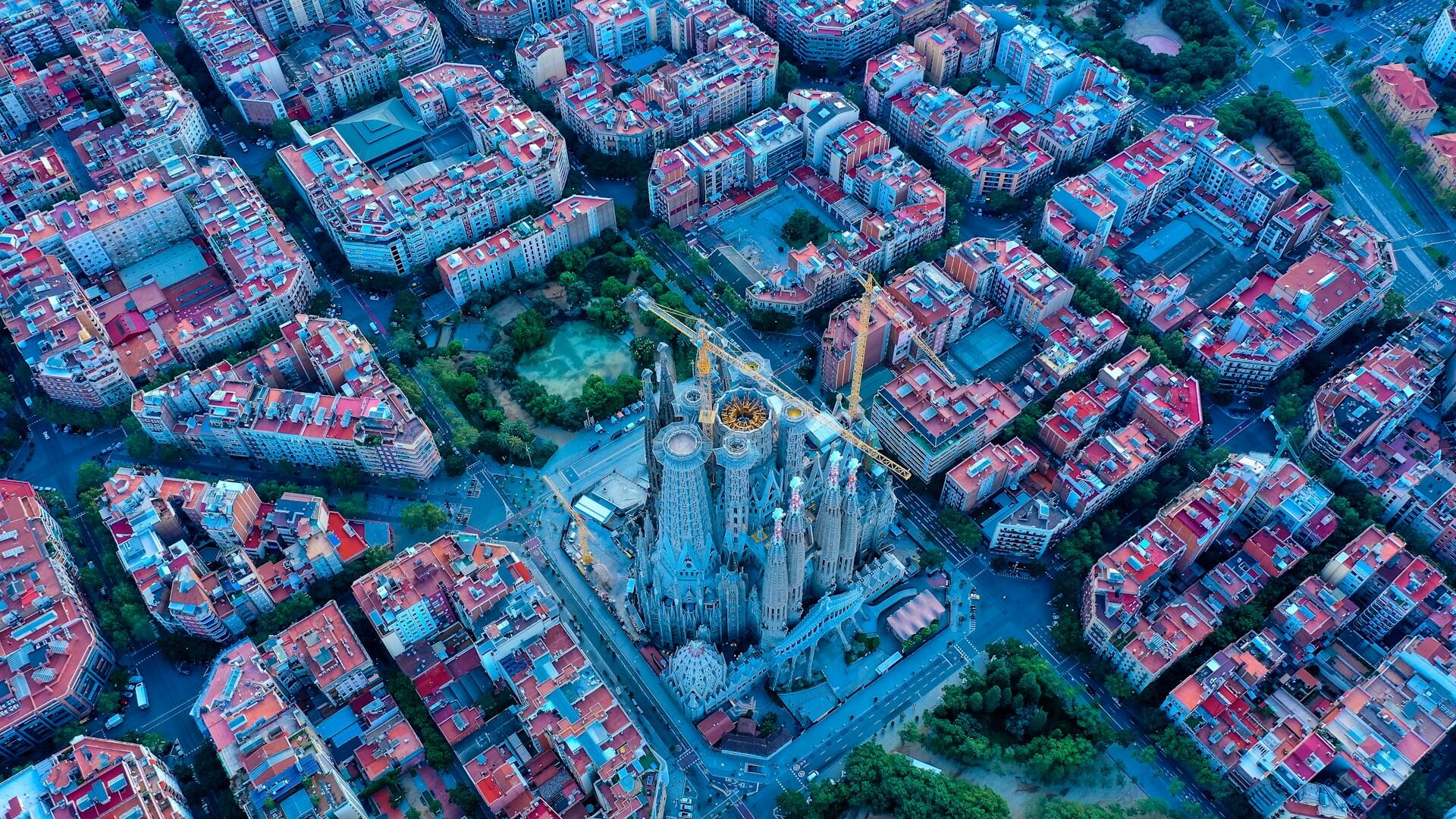 Vista aérea de la Sagrada Familia de Barcelona, rodeada de manzanas urbanísticas dispuestas en forma de cuadrícula, con tejados rojos y una exuberante vegetación, que muestra una intrincada obra maestra arquitectónica.