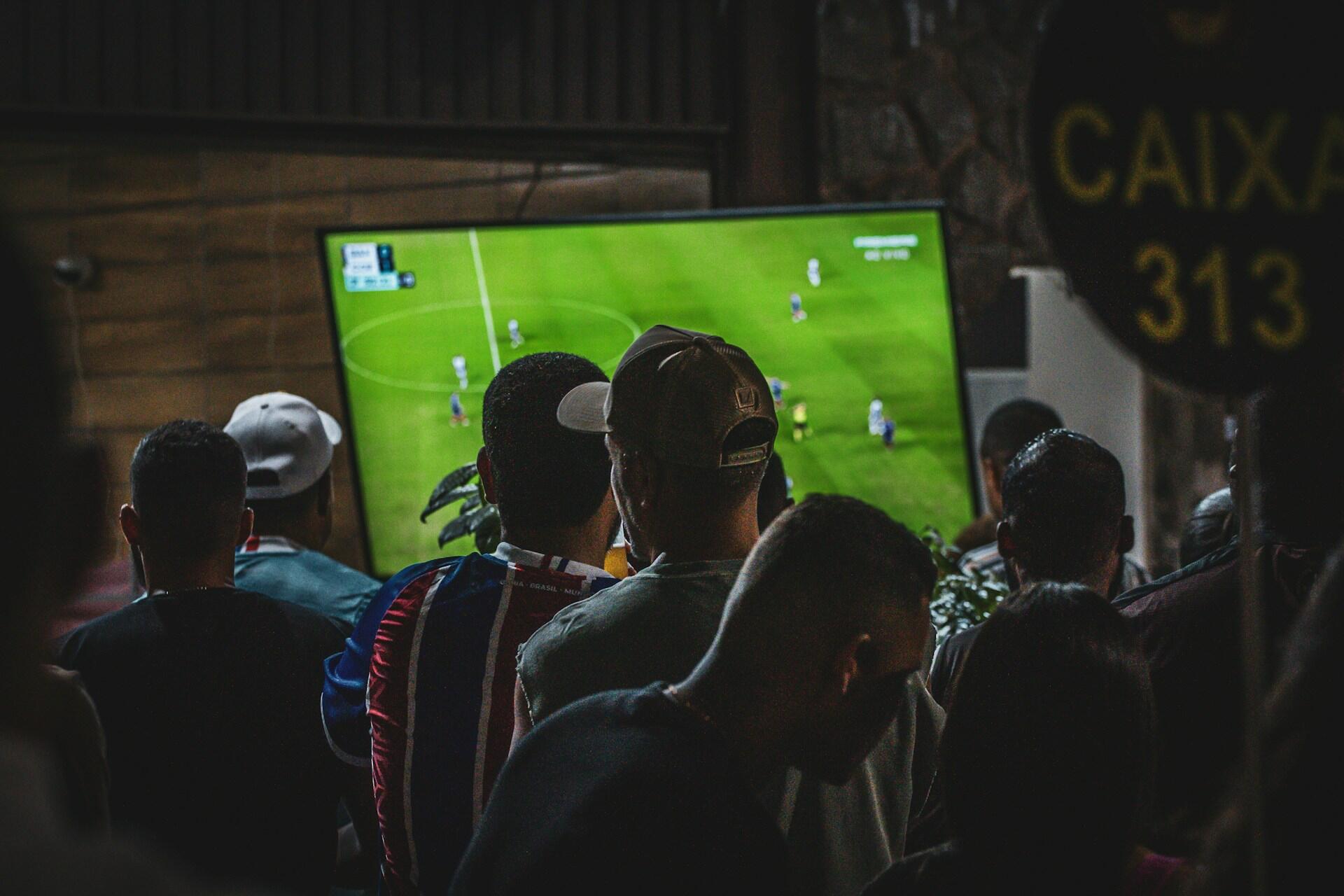 La gente se apiña para ver un partido de fútbol en una gran pantalla de televisión, en una sala con poca luz. El ambiente es tenso y de gran concentración.