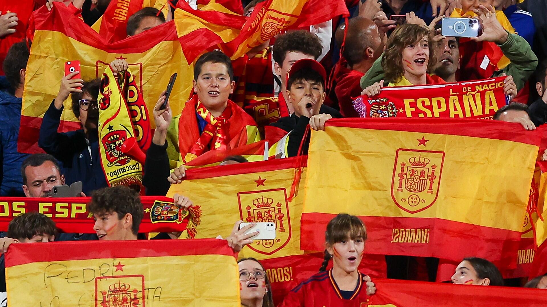 Un grupo de entusiastas aficionados al fútbol españoles que agitan banderas de vivos colores rojos y amarillos y animan con energía, reflejando la emoción del evento.