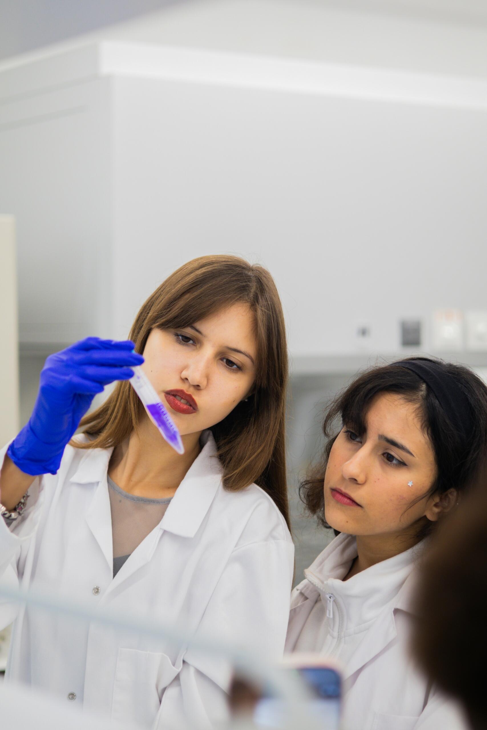 Dos científicas con bata de laboratorio y guantes examinando un tubo de ensayo en un laboratorio.